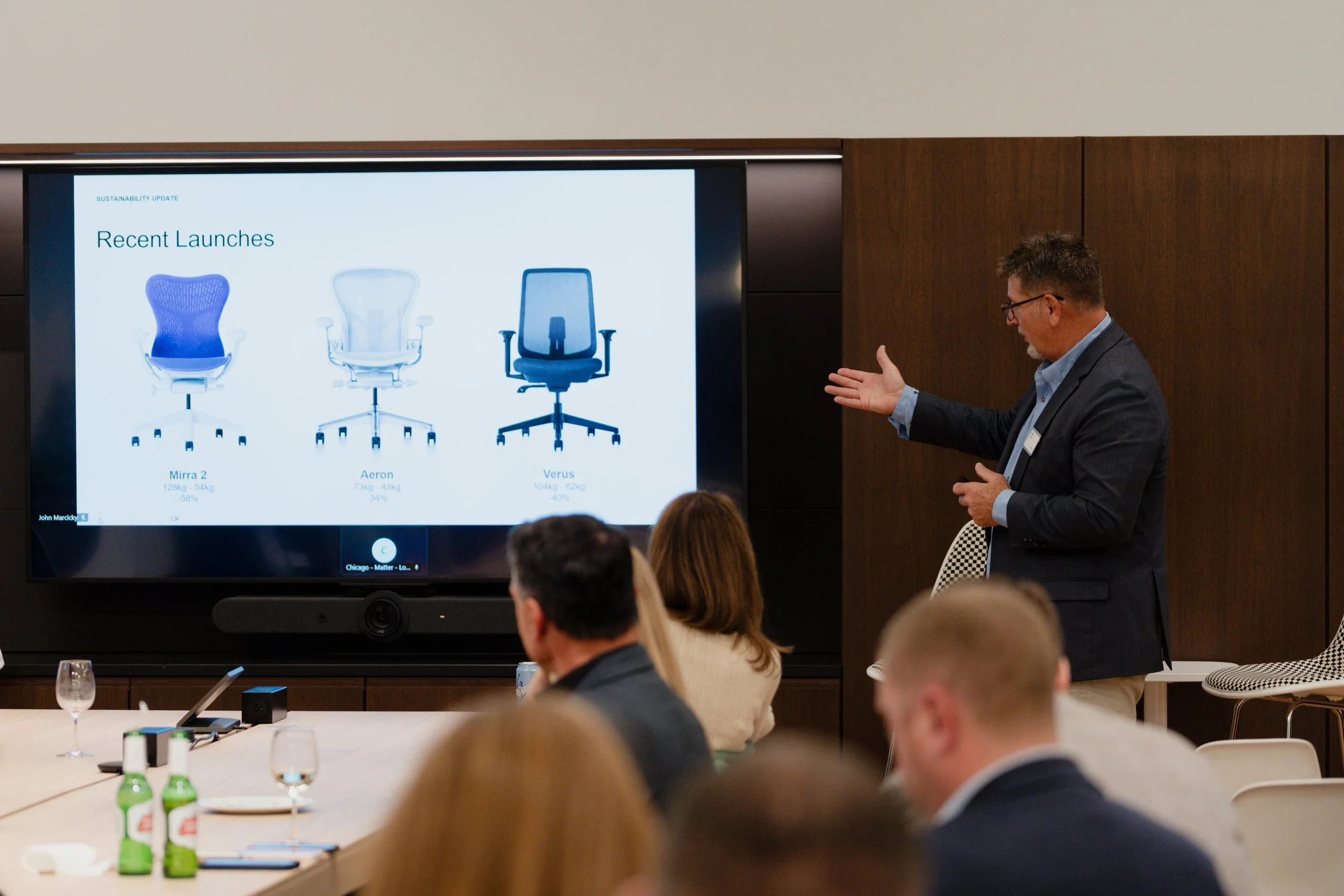 Business presentation in a conference room showing three recent chair models on a screen labeled Mirra 2, Aeron, and Verus, with a man pointing at the screen and an audience seated at a table.