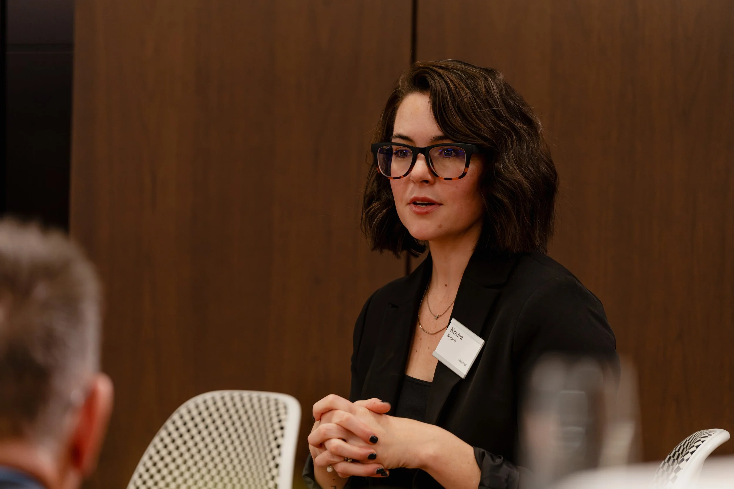 Woman speaking during a professional meeting or event, wearing glasses, a black blazer, and a name tag, with brown hair and a neutral background.