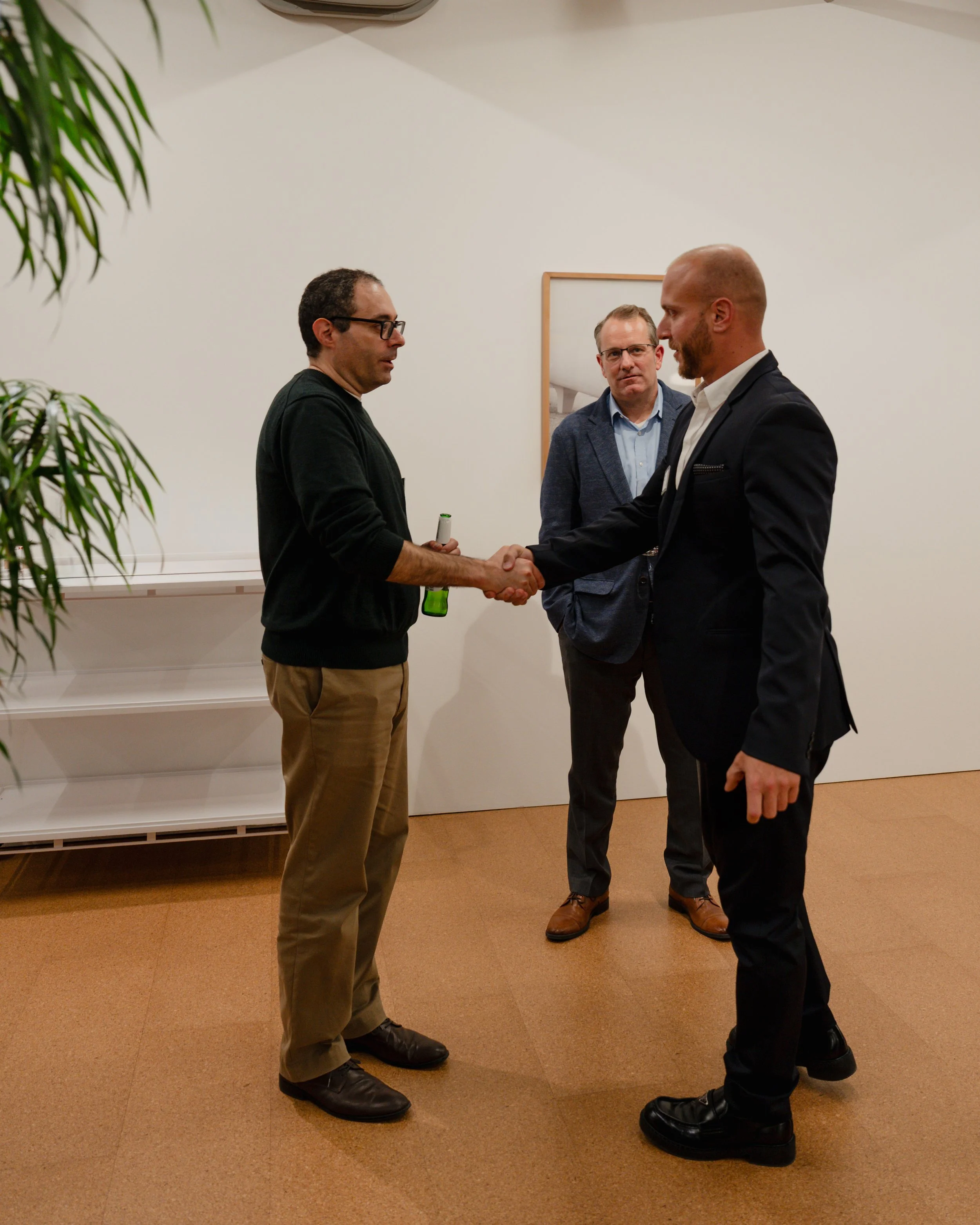 Two men shaking hands while a third man looks on in a professional setting, with a beige carpet and white walls.