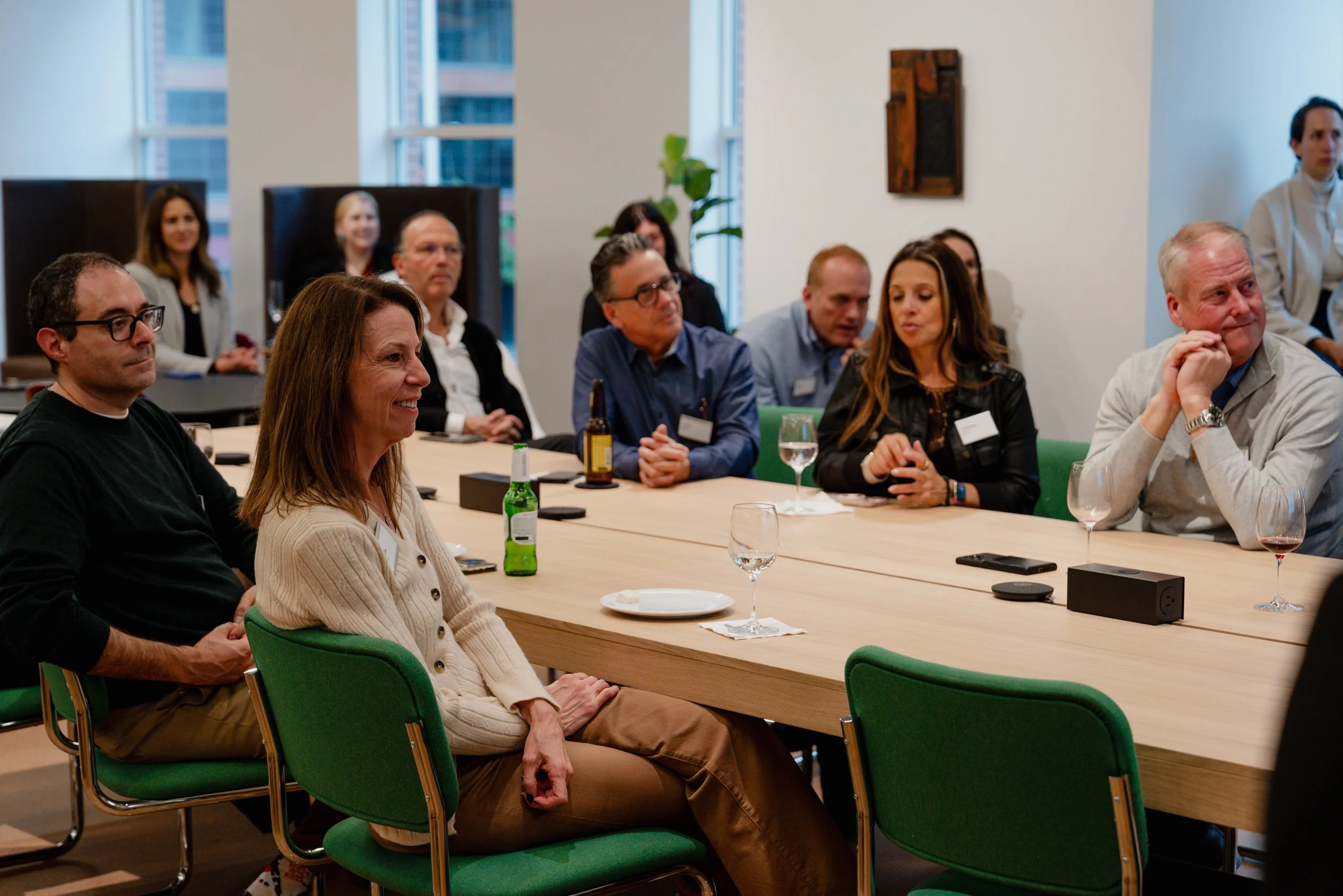 People sitting around a conference table in a modern office, attentively listening to a speaker, with drinks and electronic devices on the table.
