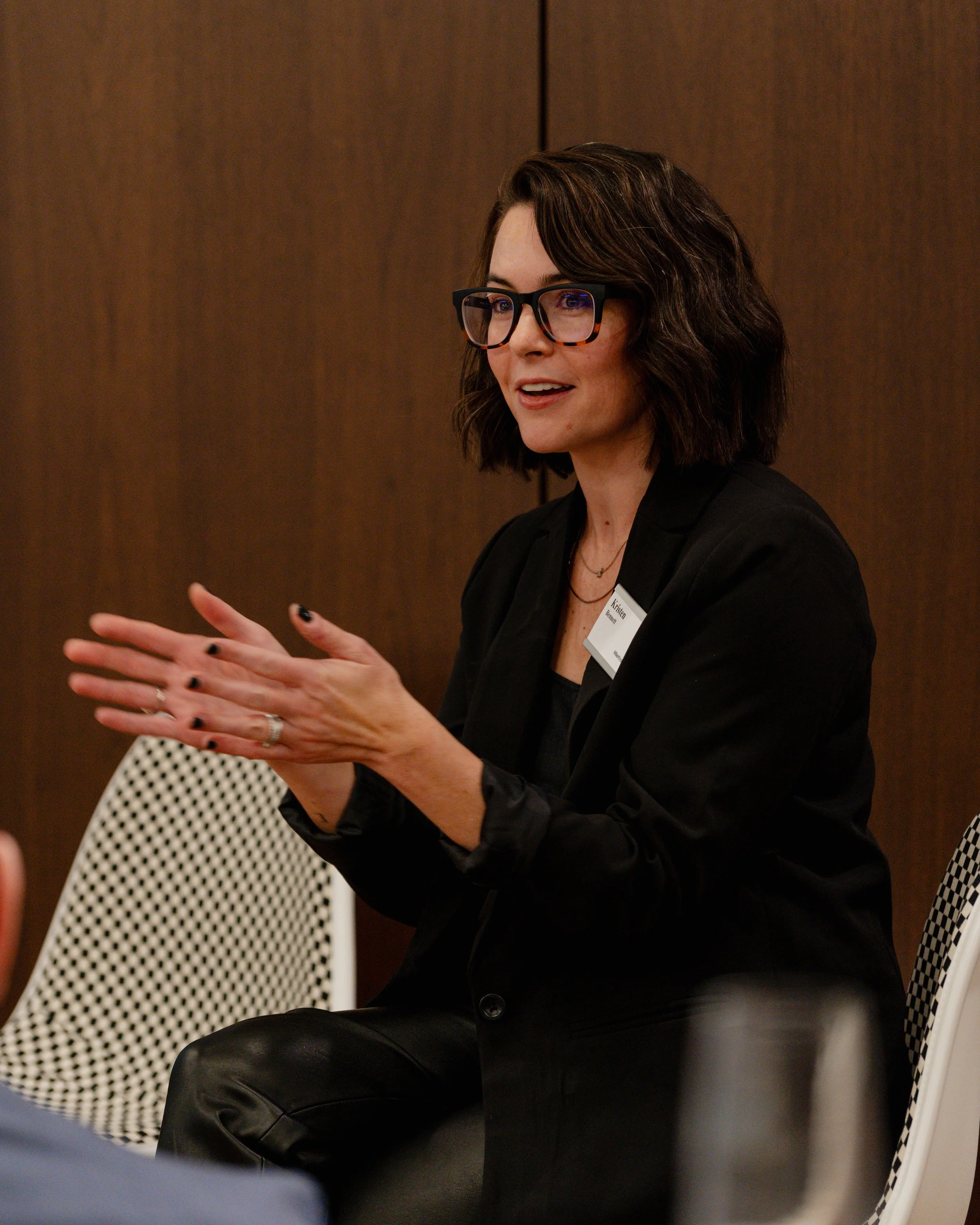 A woman with dark wavy hair, glasses, and a black blazer speaking and gesturing with her hands during a discussion.