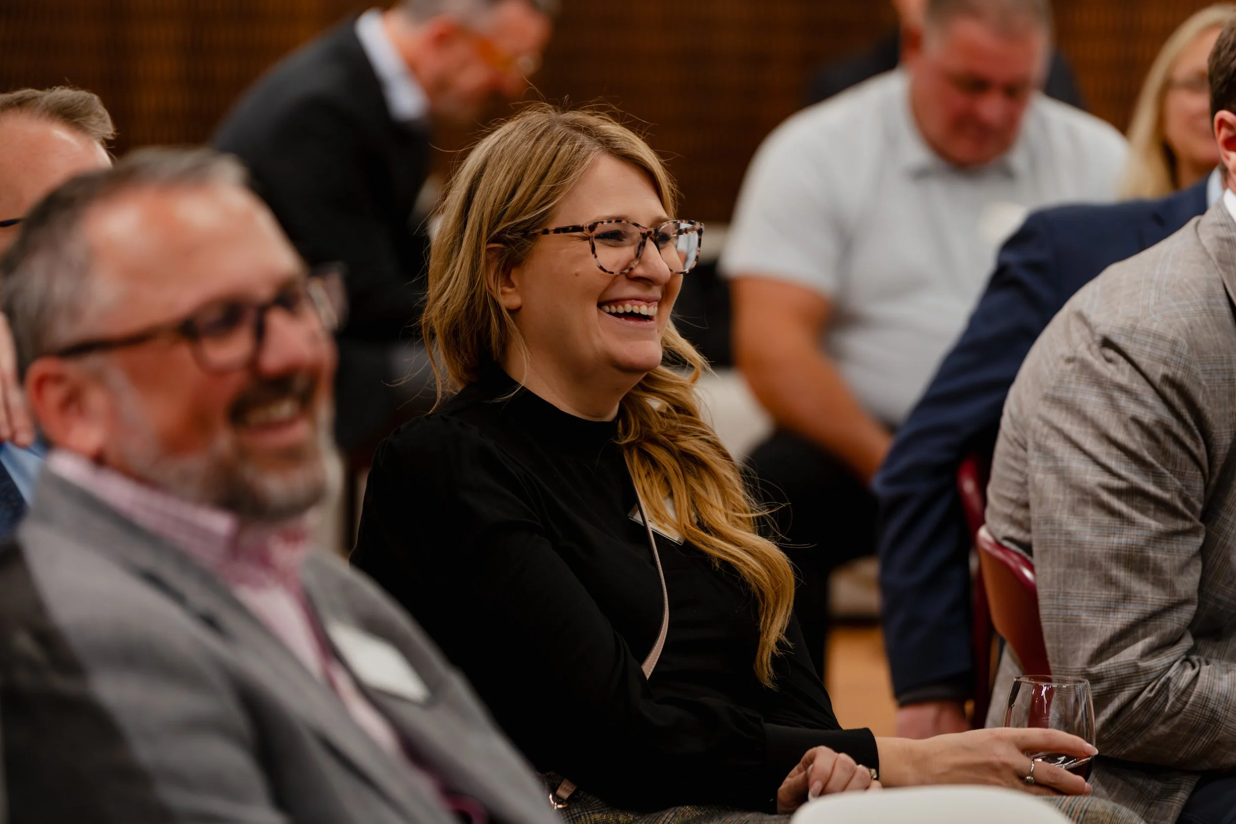 A woman with long reddish hair and glasses smiling at a conference or event, holding a glass of wine.