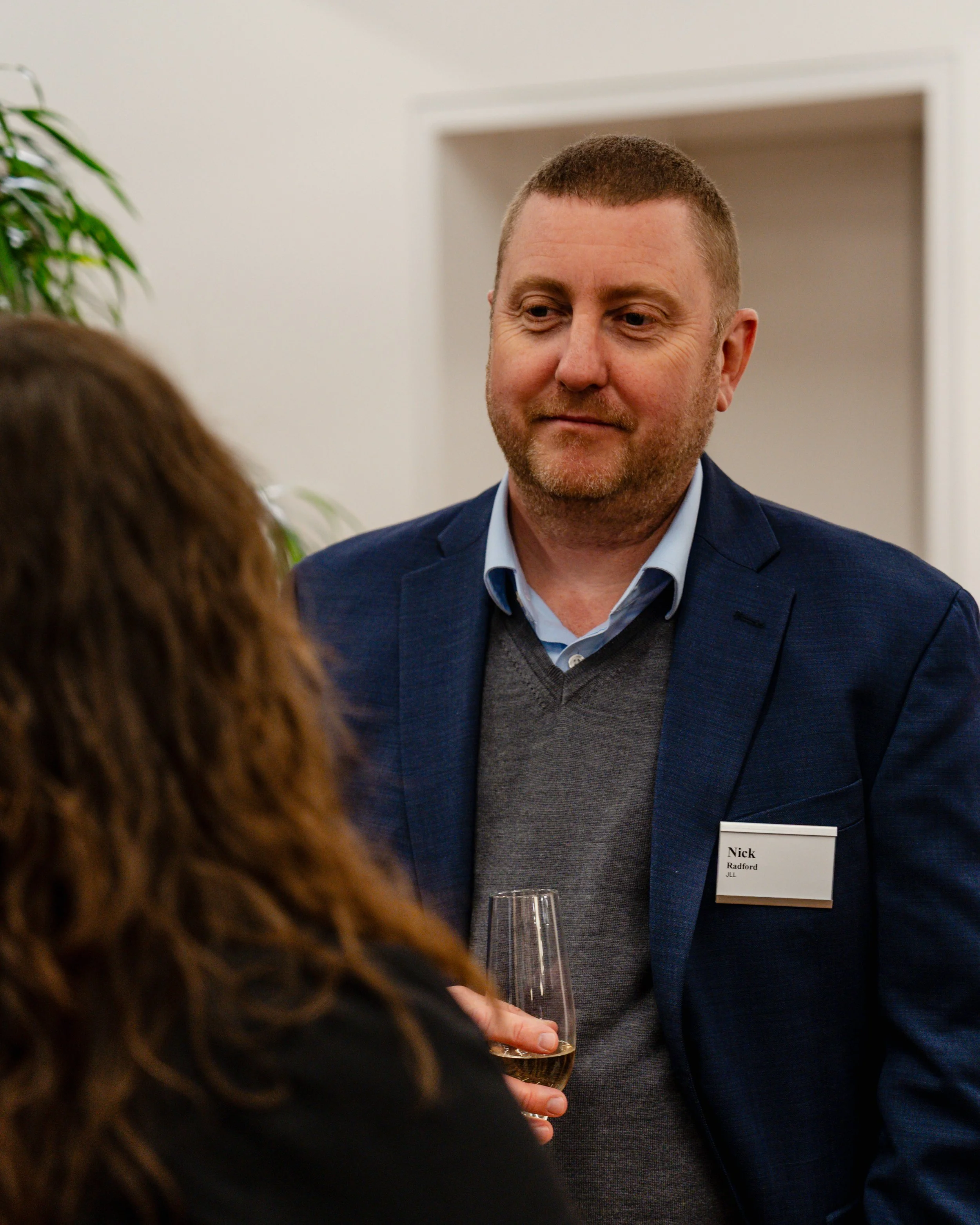 A man in a blue blazer and gray sweater holding a glass of white wine, engaging in conversation with a woman whose back is turned to the camera, at a professional networking event.