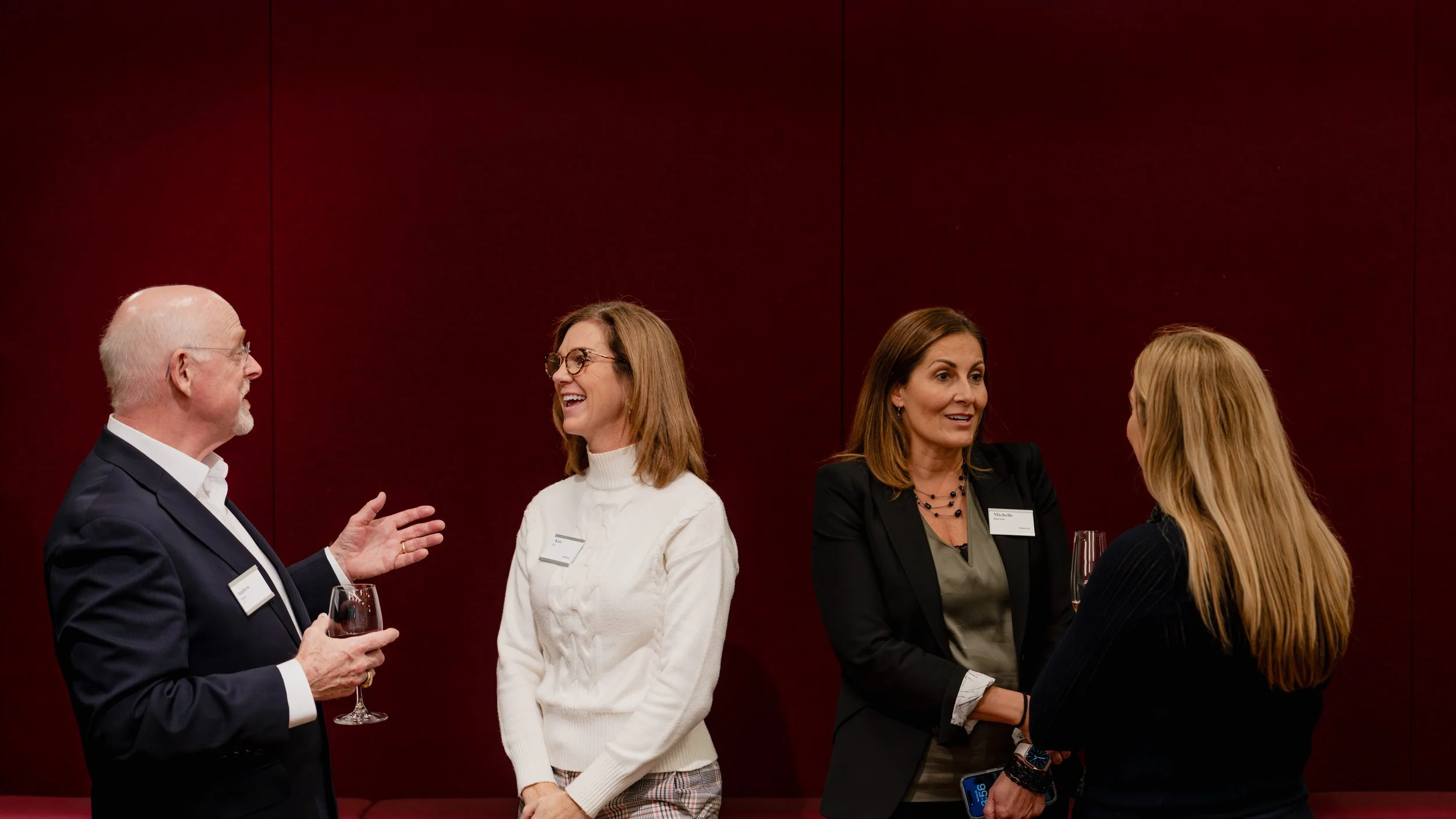 Four adults, two men and two women, engaged in conversation at a professional networking event, standing against a plain dark red wall.