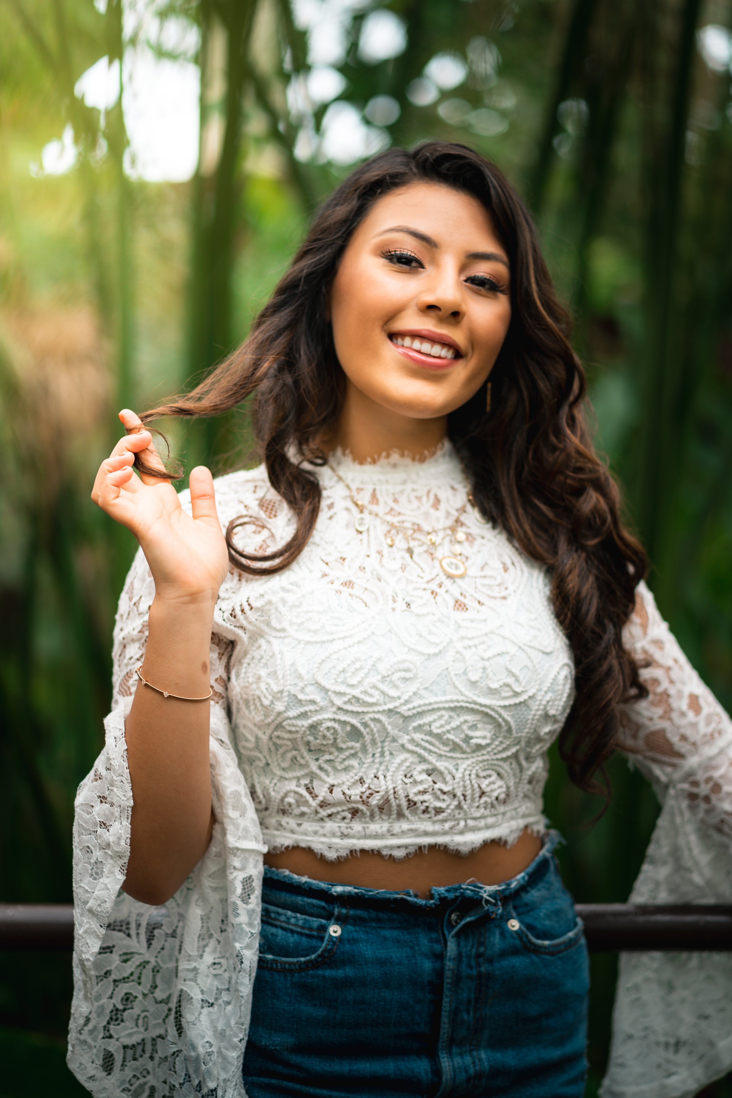 A portrait of a woman with long dark curly hair smiling, holding a strand of her hair, in a green outdoor setting with blurred foliage in the background.