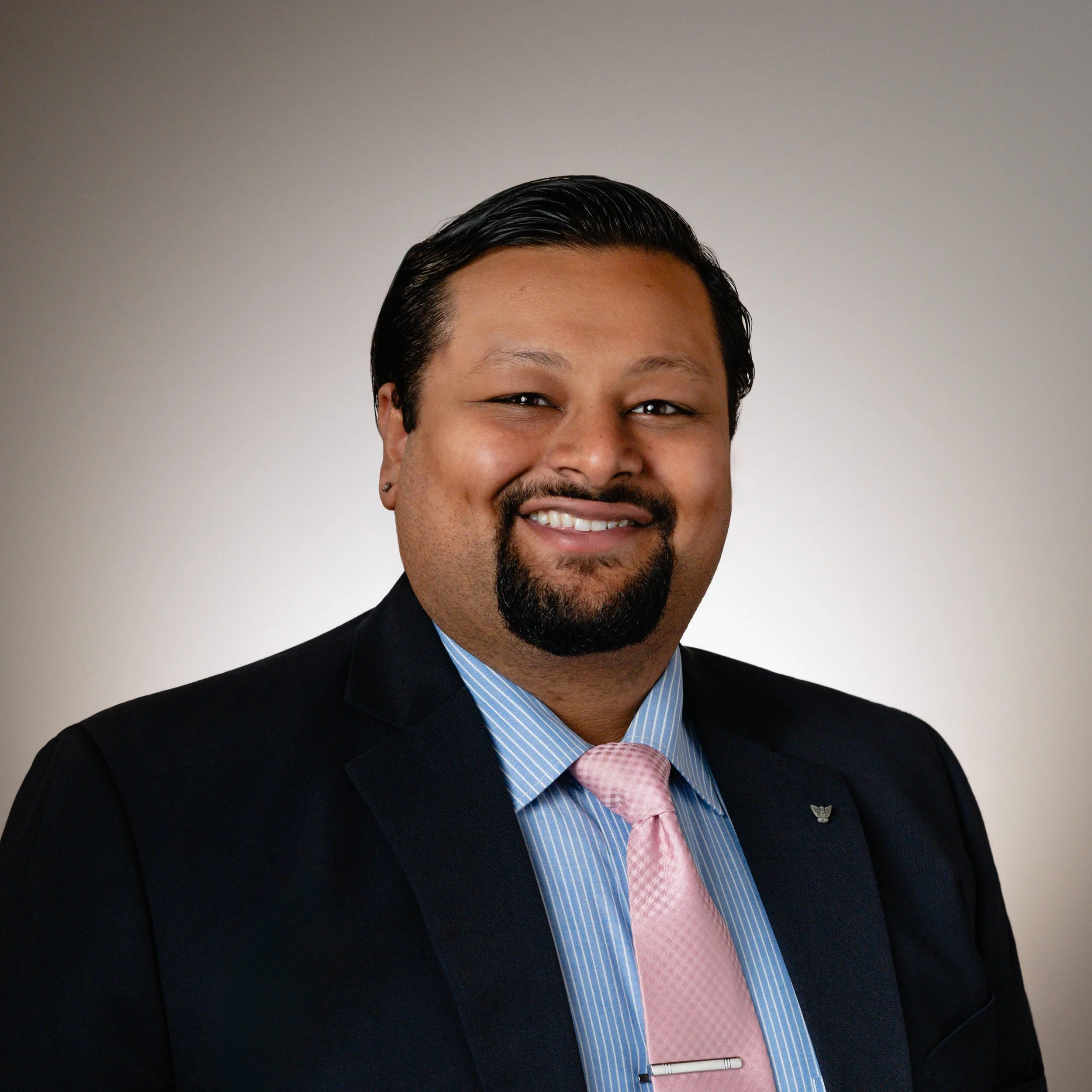 Professional headshot of a smiling man with black hair, a goatee, wearing a black suit, blue striped shirt, and pink tie, against a plain gray background.