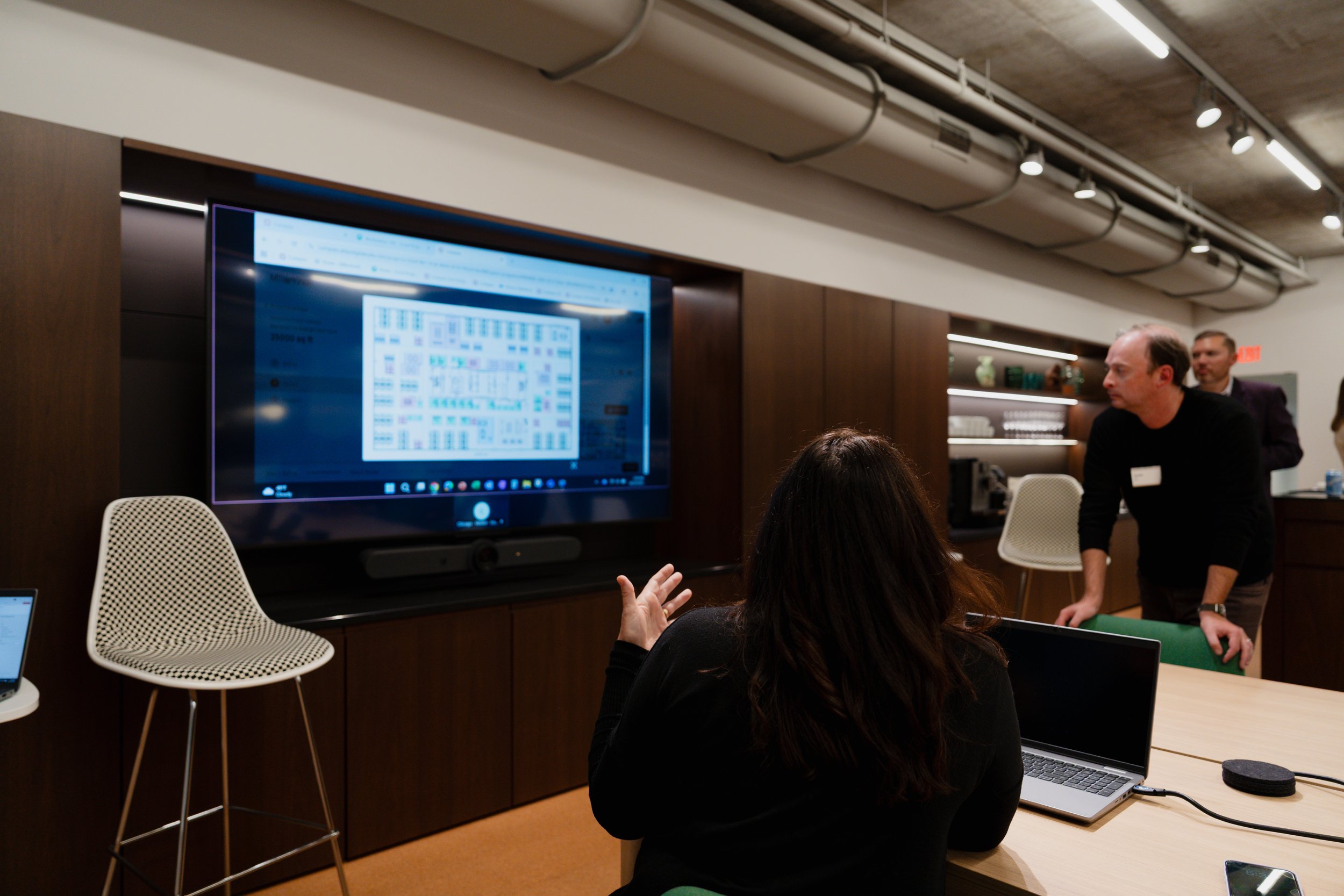 A woman sitting at a conference table with a laptop, raising her hand, in a room with a large screen displaying a spreadsheet or similar document. Two men are standing nearby, engaging in conversation.