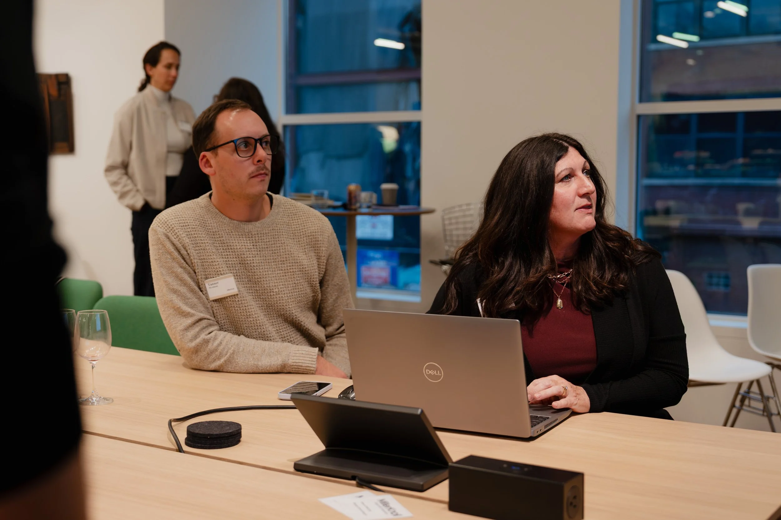 Two women sitting at a conference table, one with a Dell laptop, in a meeting room with large windows. Other people are standing in the background.
