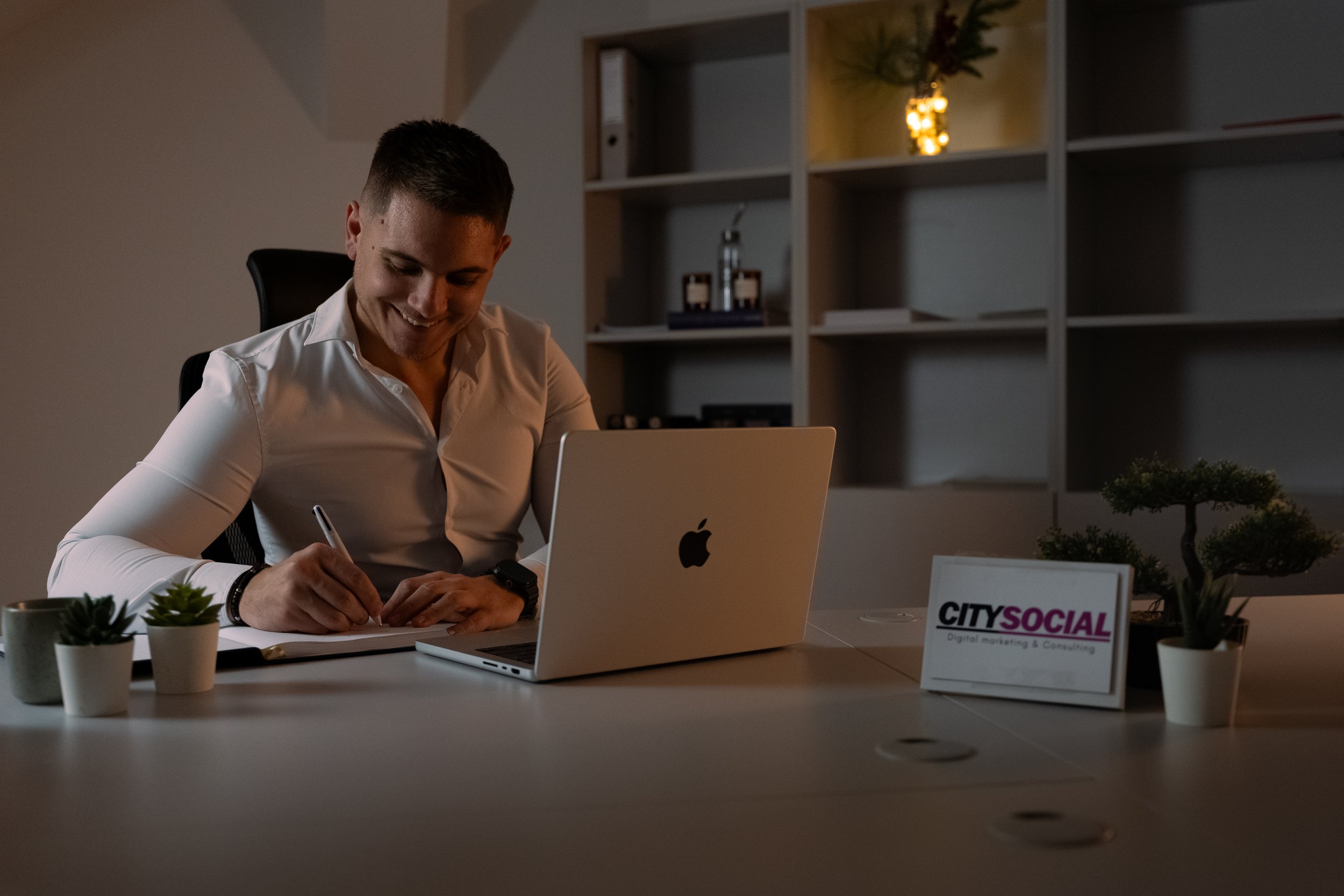 A man sitting at a desk in an office, smiling and writing in a notebook, with a silver MacBook and potted plants on the desk; a sign that reads 'CITY SOCIAL' and shelves with decorative items in the background.