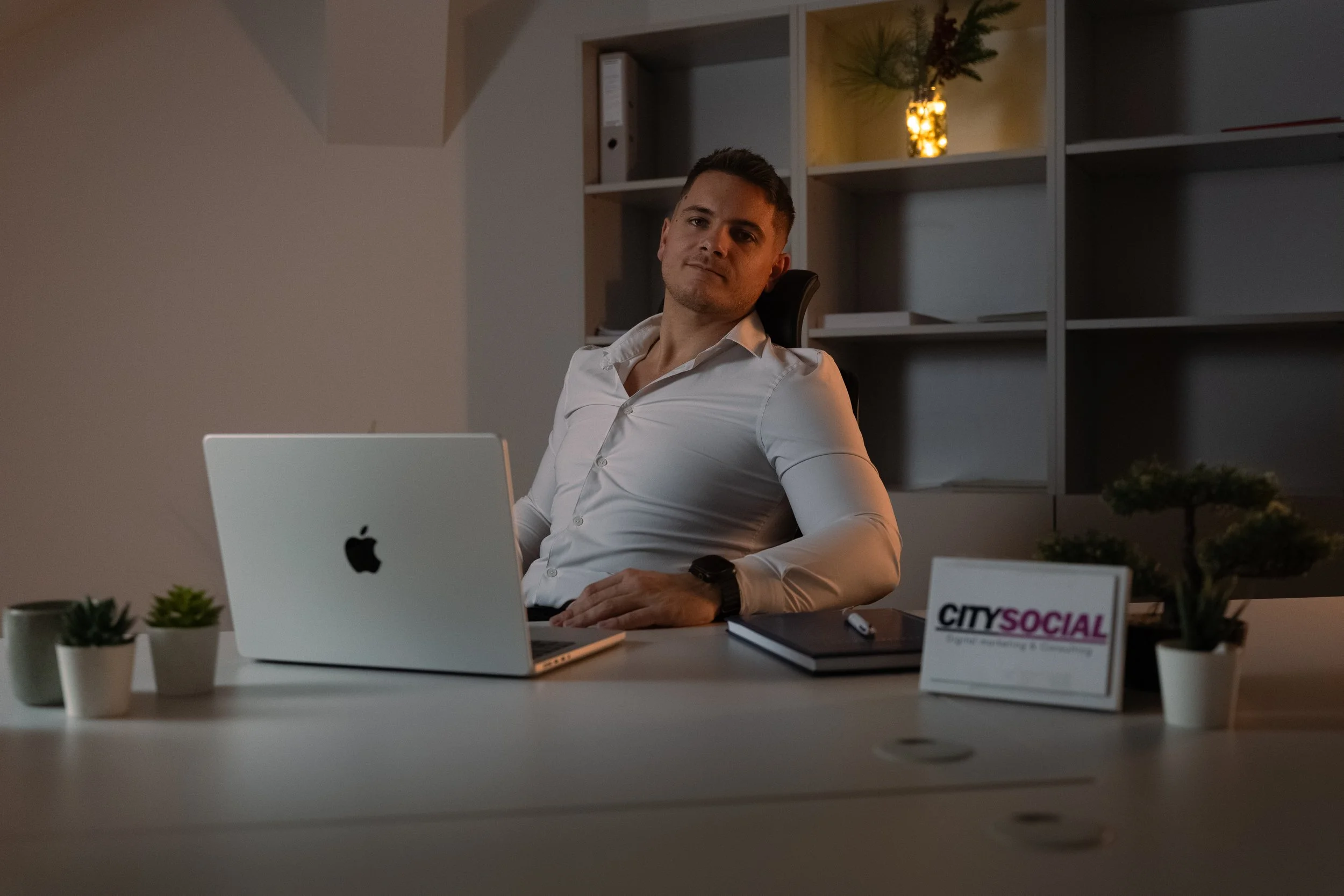 Man sitting at a desk in an office, with a laptop, potted plants, a tablet, and a sign that reads 'City Social' under dim lighting.