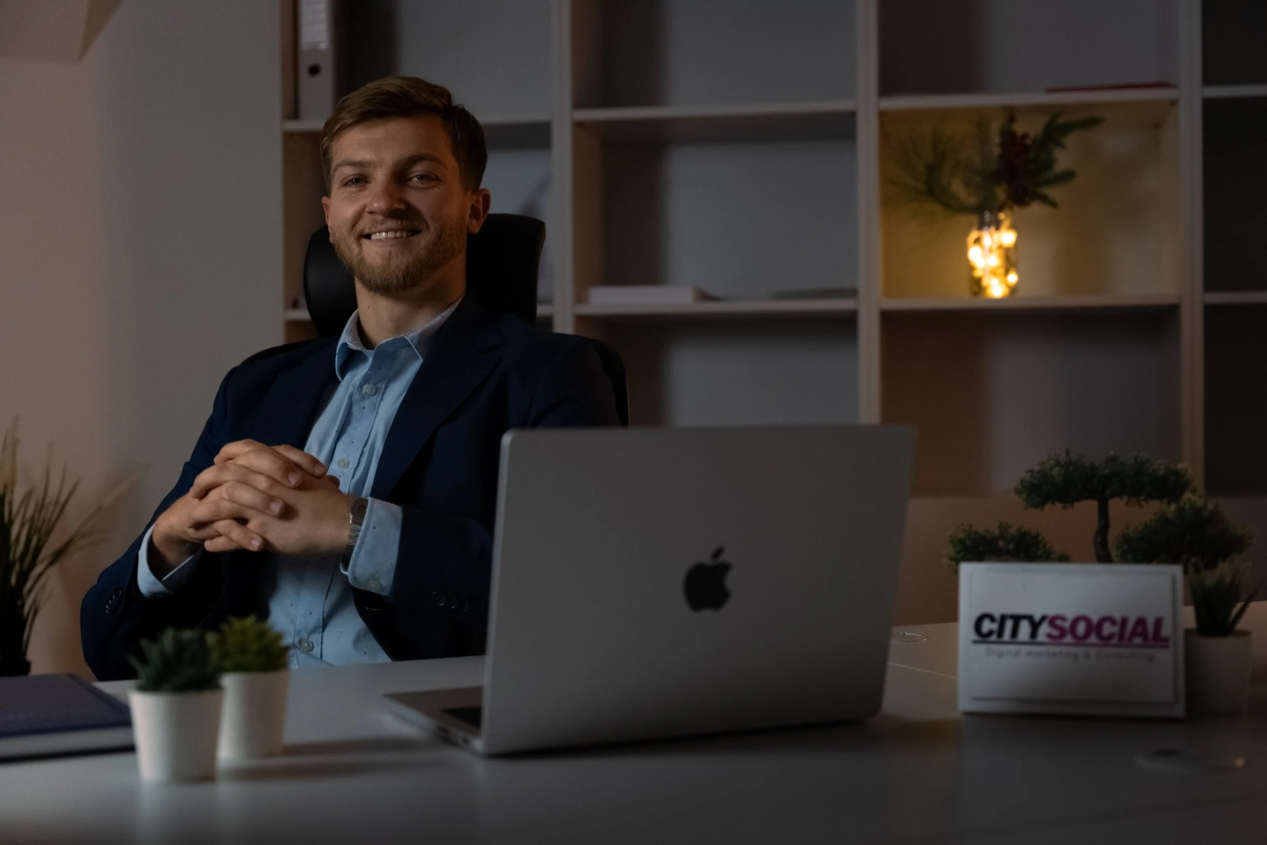 A man with a beard and smiling face sitting at a desk in an office, wearing a dark suit jacket and light blue shirt. The desk has a silver Apple MacBook, a small potted plant, a notebook, and a sign that reads 'CITY SOCIAL Digital marketing & consulting'. The background features shelving with a decorative plant and a lit vase.