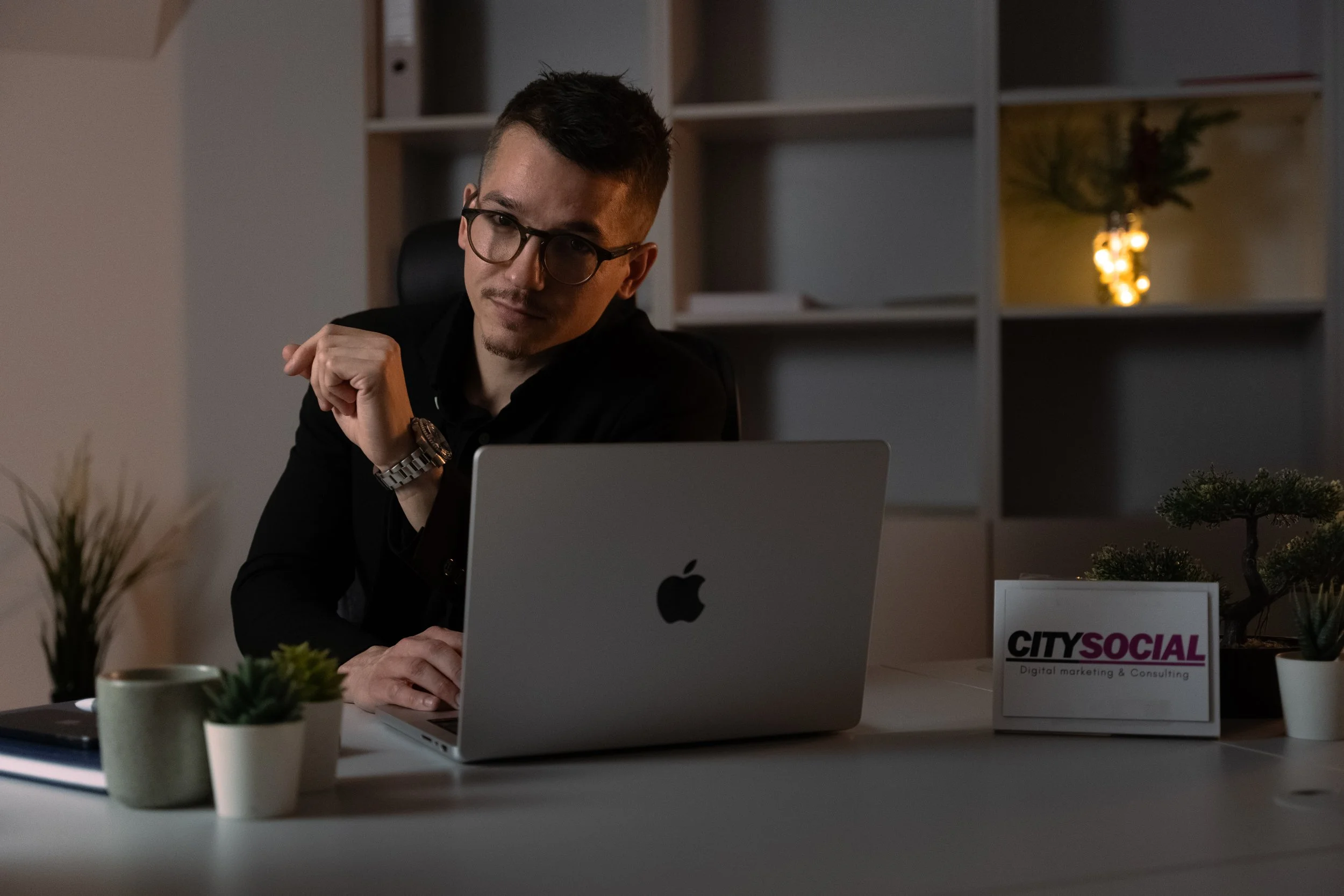 A man with glasses and a watch sits at a desk in front of an open MacBook laptop, with plants and a sign that reads 'CITY SOCIAL Digital marketing & Consulting' in an office with shelves and a lit decorative plant in the background.