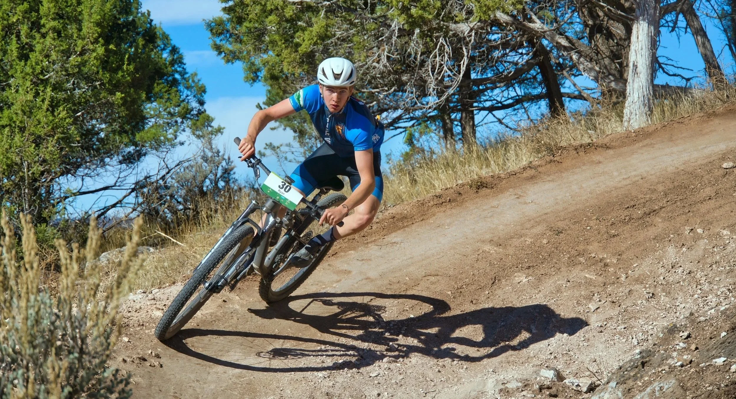 Young male mountain biker in blue jersey and helmet riding on a dirt trail through a wooded area with trees in the background.