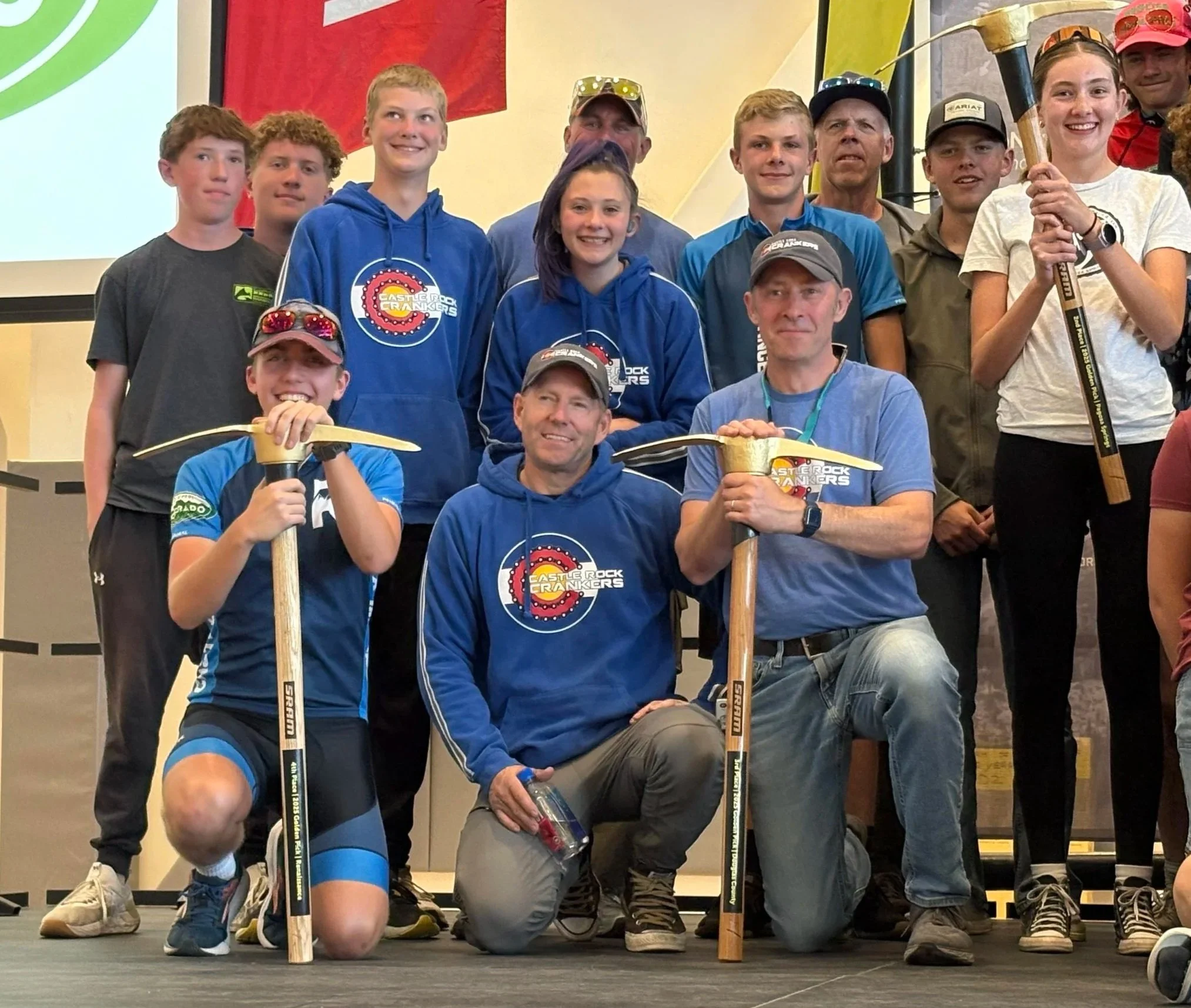 Group of young people and adults on stage, some holding rock hammers, at a rock climbing or caving event, with a large screen displaying a logo in the background.