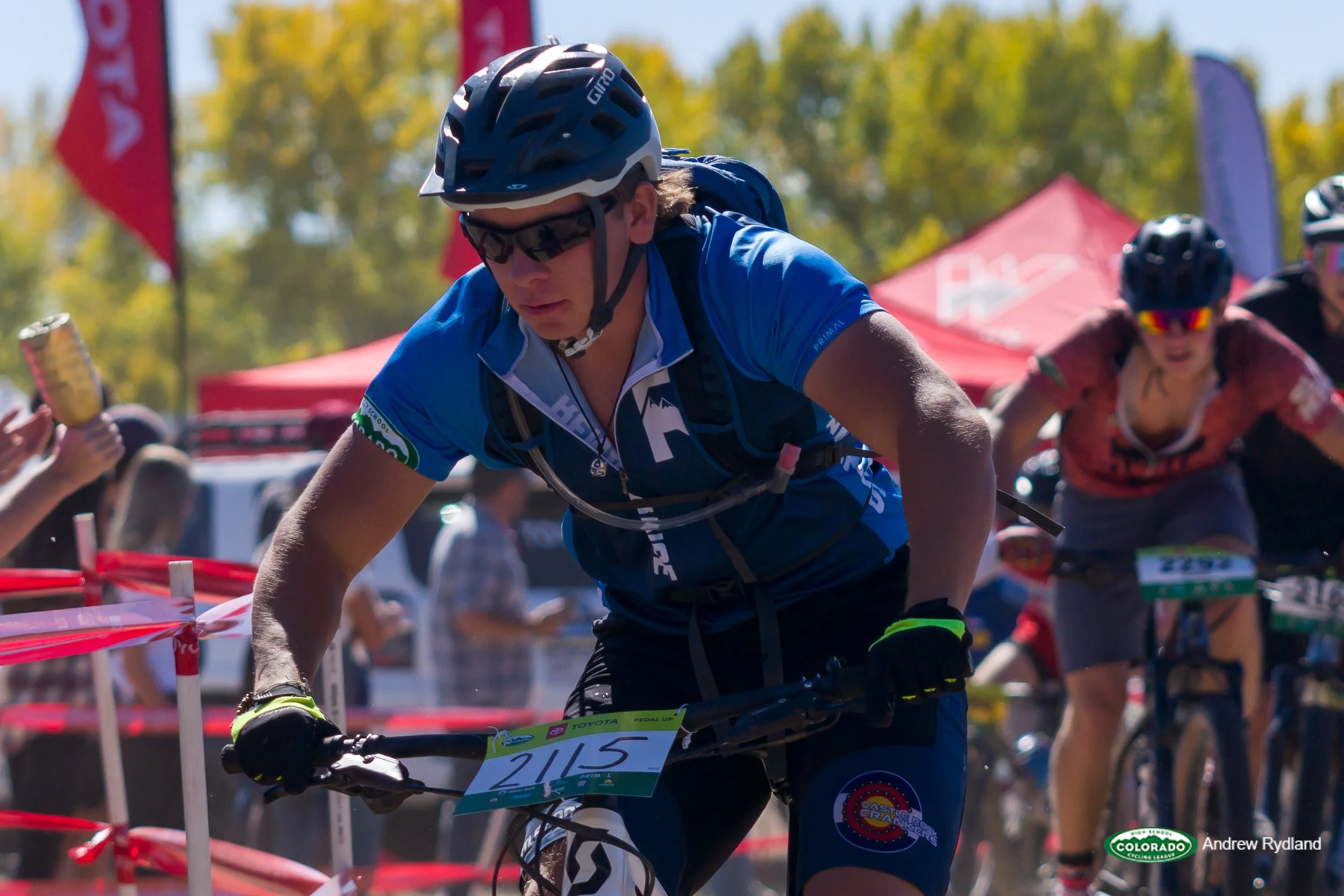A male cyclist wearing a blue jersey, helmet, and sunglasses rides a mountain bike during a race, with other cyclists and spectators in the background.
