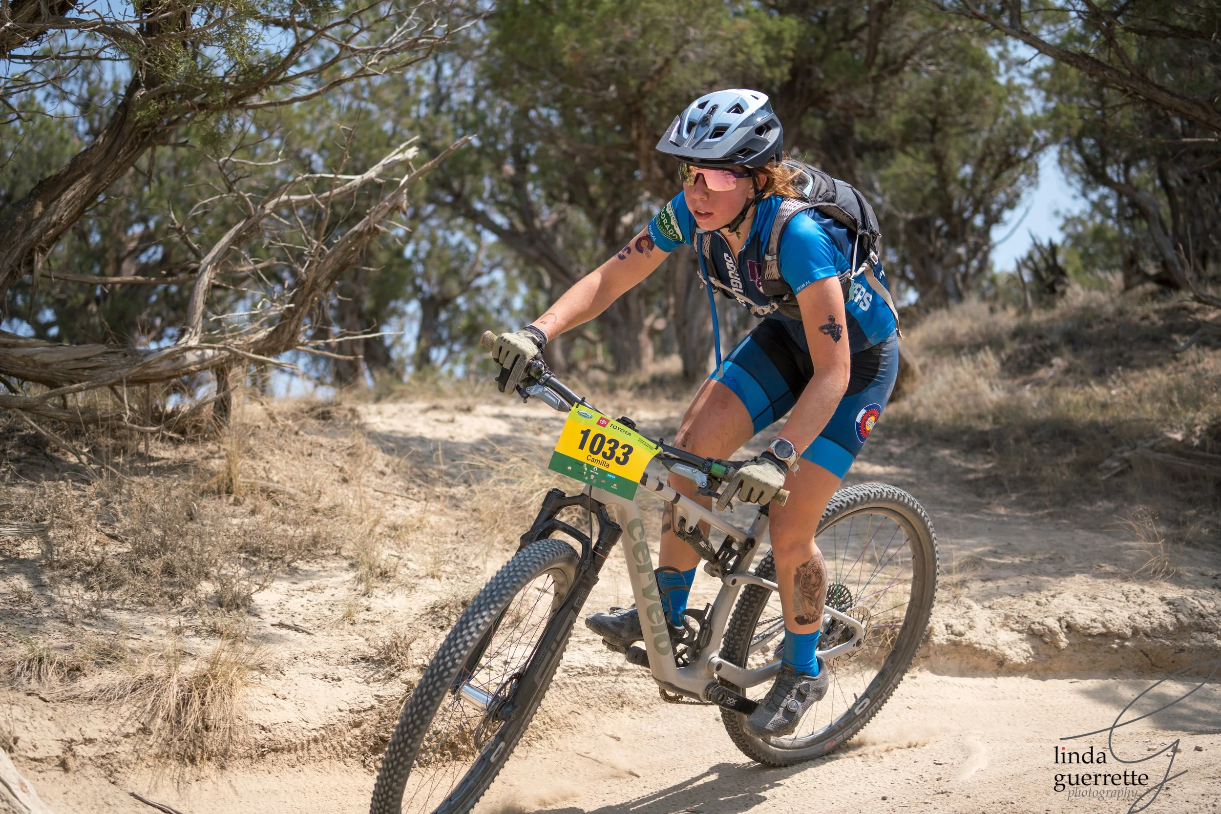 A female mountain biker in blue gear riding on a sandy trail through trees, wearing a helmet, sunglasses, and a backpack, with a yellow race bib numbered 1033.