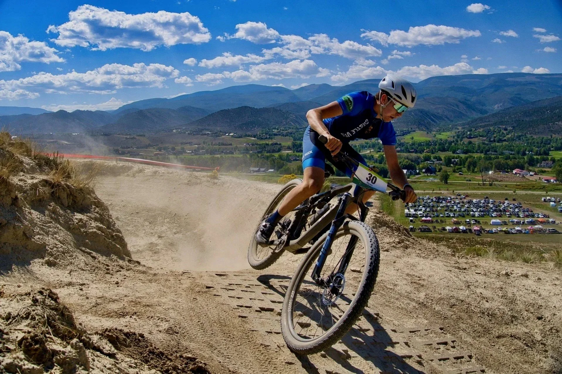 A mountain biker in blue gear and a white helmet leans into a turn on a dirt trail with a mountainous landscape and blue sky in the background.