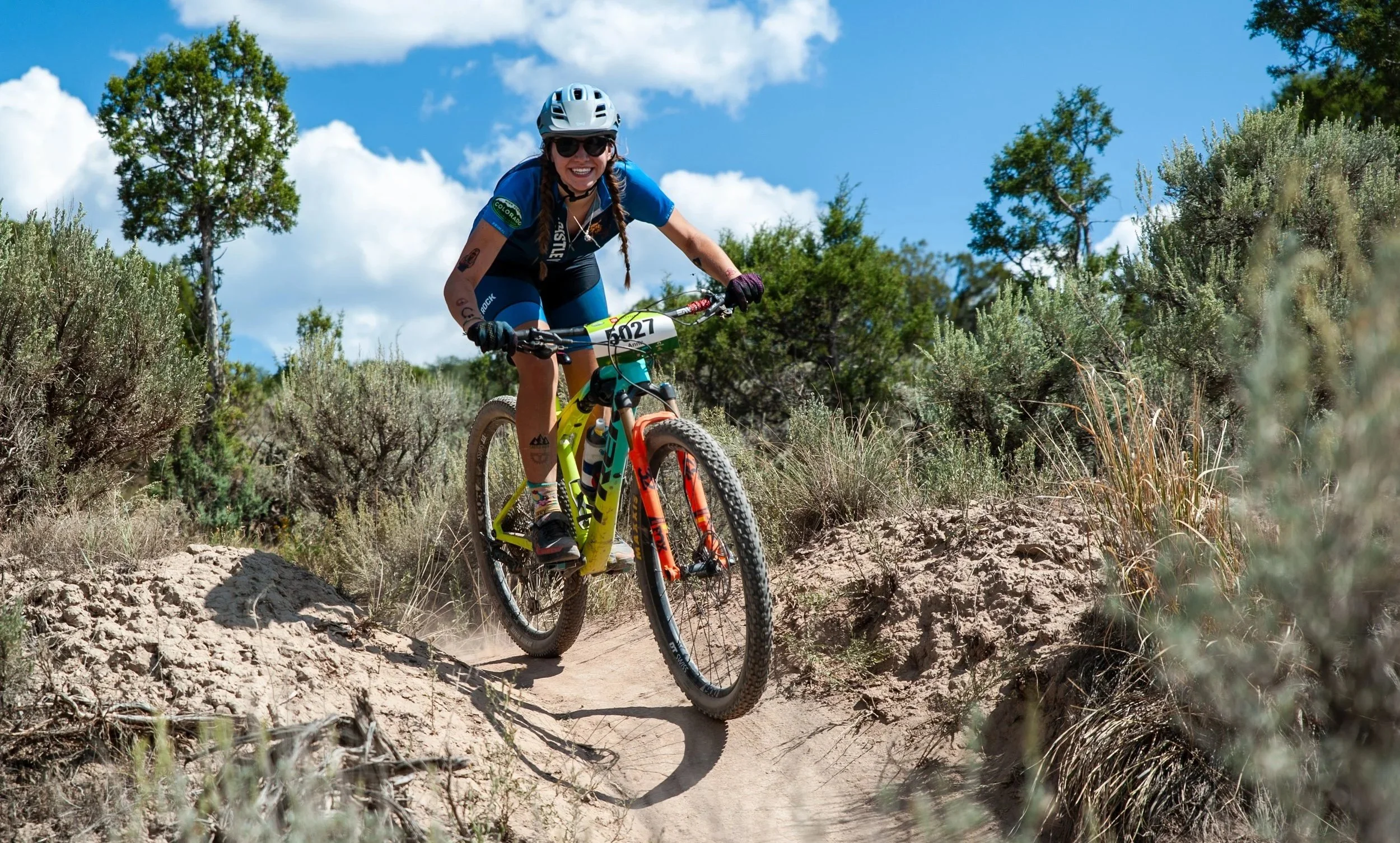A woman riding a mountain bike on a dirt trail surrounded by bushes and trees, wearing a helmet, sunglasses, and athletic clothing, smiling.