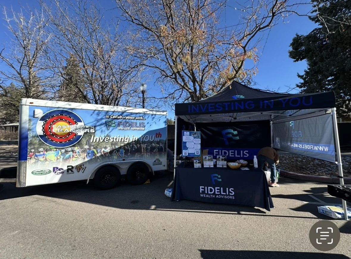 A promotional setup for Fidellis Wealth Advisors, including a trailer and a tent with the slogan "Investing in You." The trailer features a logo of the East Lake Rockers Cycling Team, indicating collaboration. The tent displays the Fidellis logo and branding, with informational materials and a bowl on the table.
