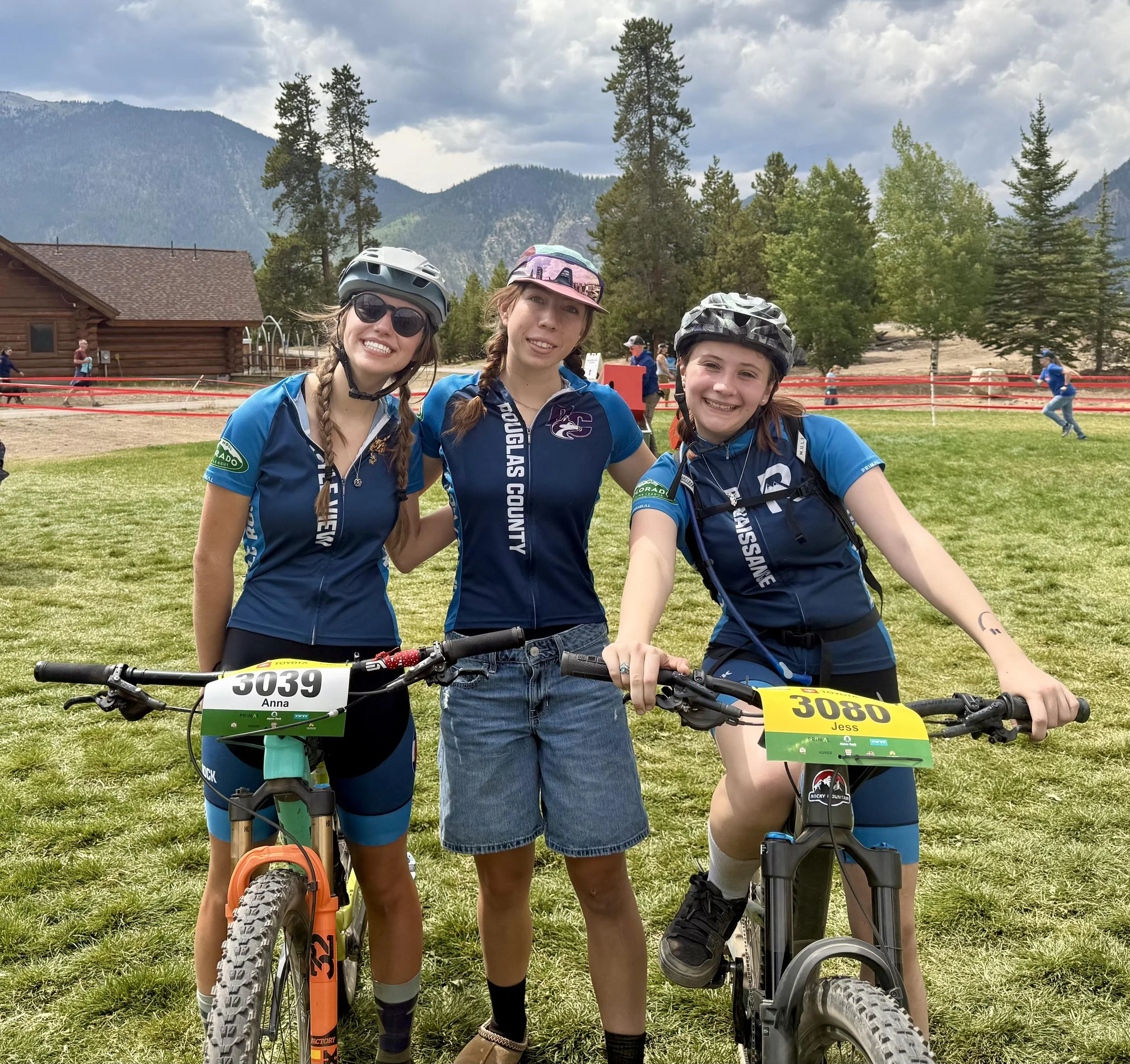 Three young women in cycling gear standing outdoors with two mountain bikes, smiling, with mountains and trees in the background.