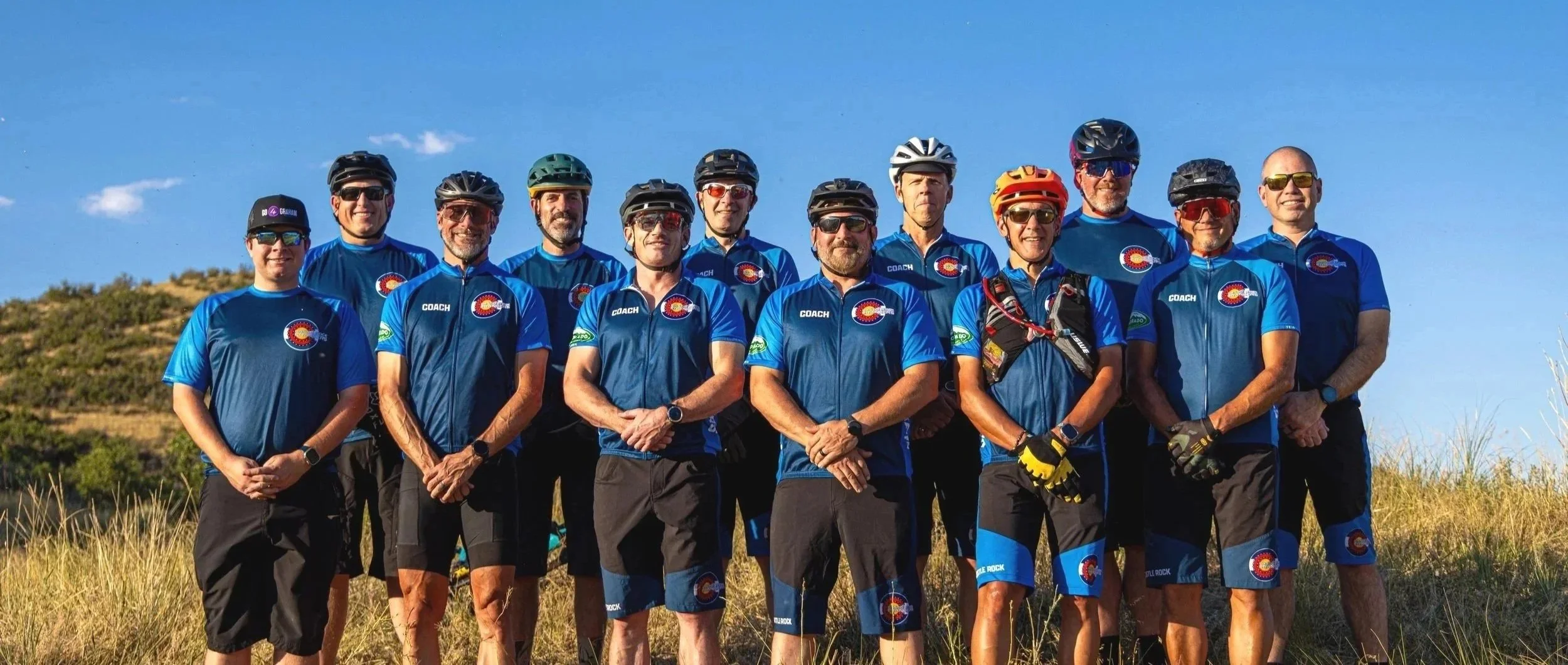 A group of fourteen cyclists standing outdoors on a grassy field, wearing matching blue cycling jerseys with Colorado Rockies logo and helmets, with clear blue sky and hills in the background.