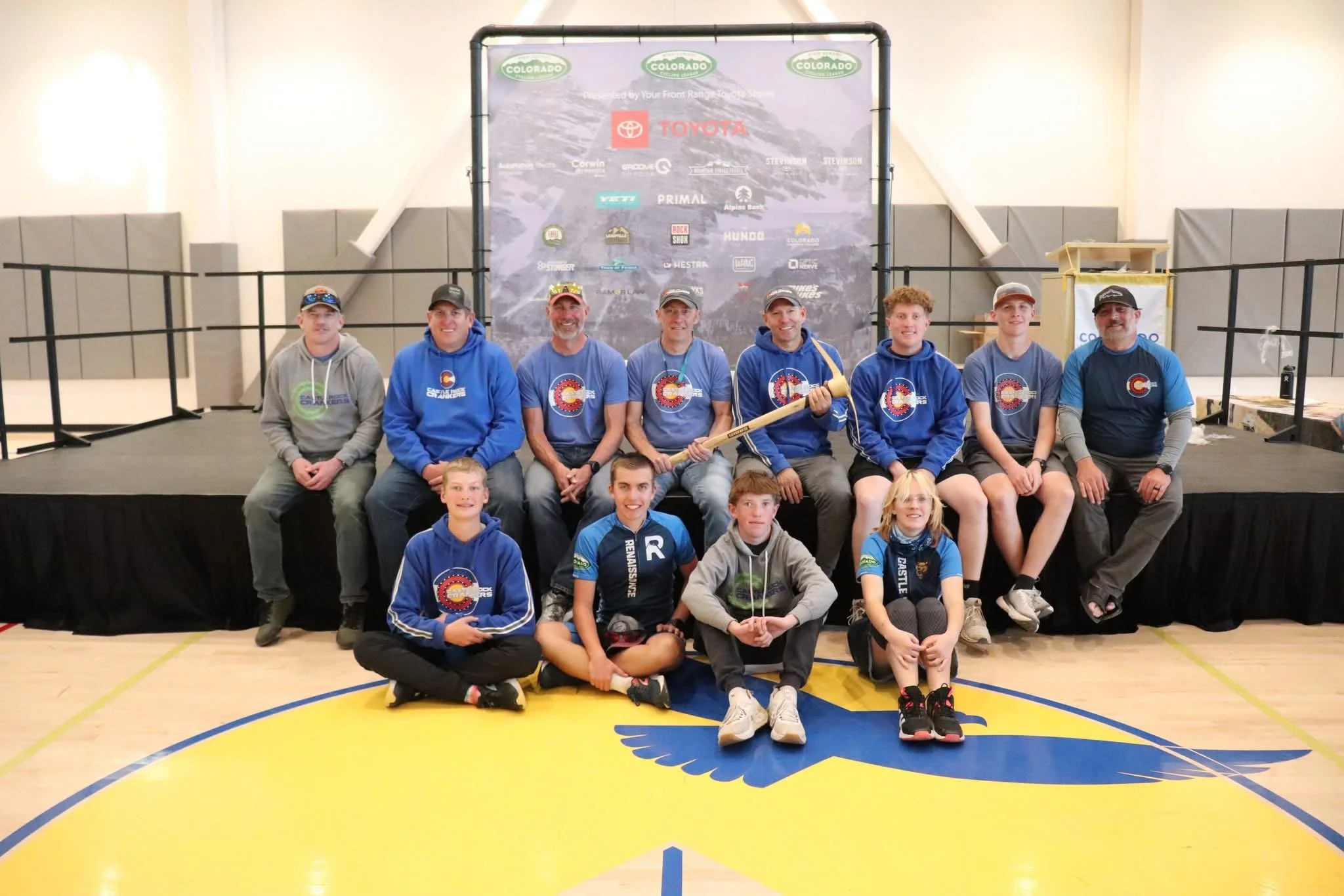 Group of people, both adults and children, sitting and standing on a stage in front of a Colorado-themed backdrop with logos, inside a gymnasium. The group appears to be at an outdoor or sports event, with some individuals wearing Colorado-themed clo