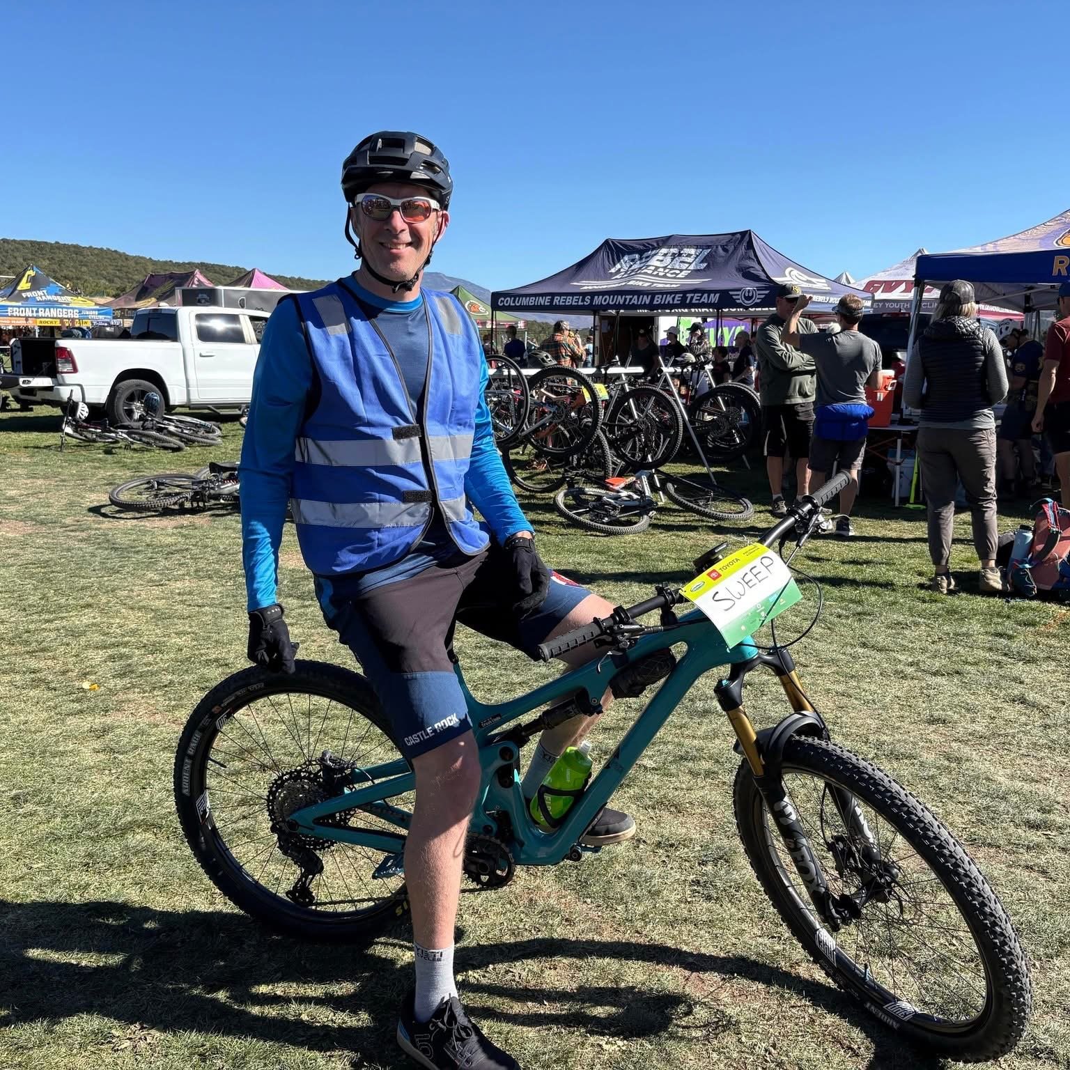 A man in cycling gear, including a helmet and sunglasses, sitting on a mountain bike labeled 'SWEEP' at an outdoor biking event with tents and people in the background.