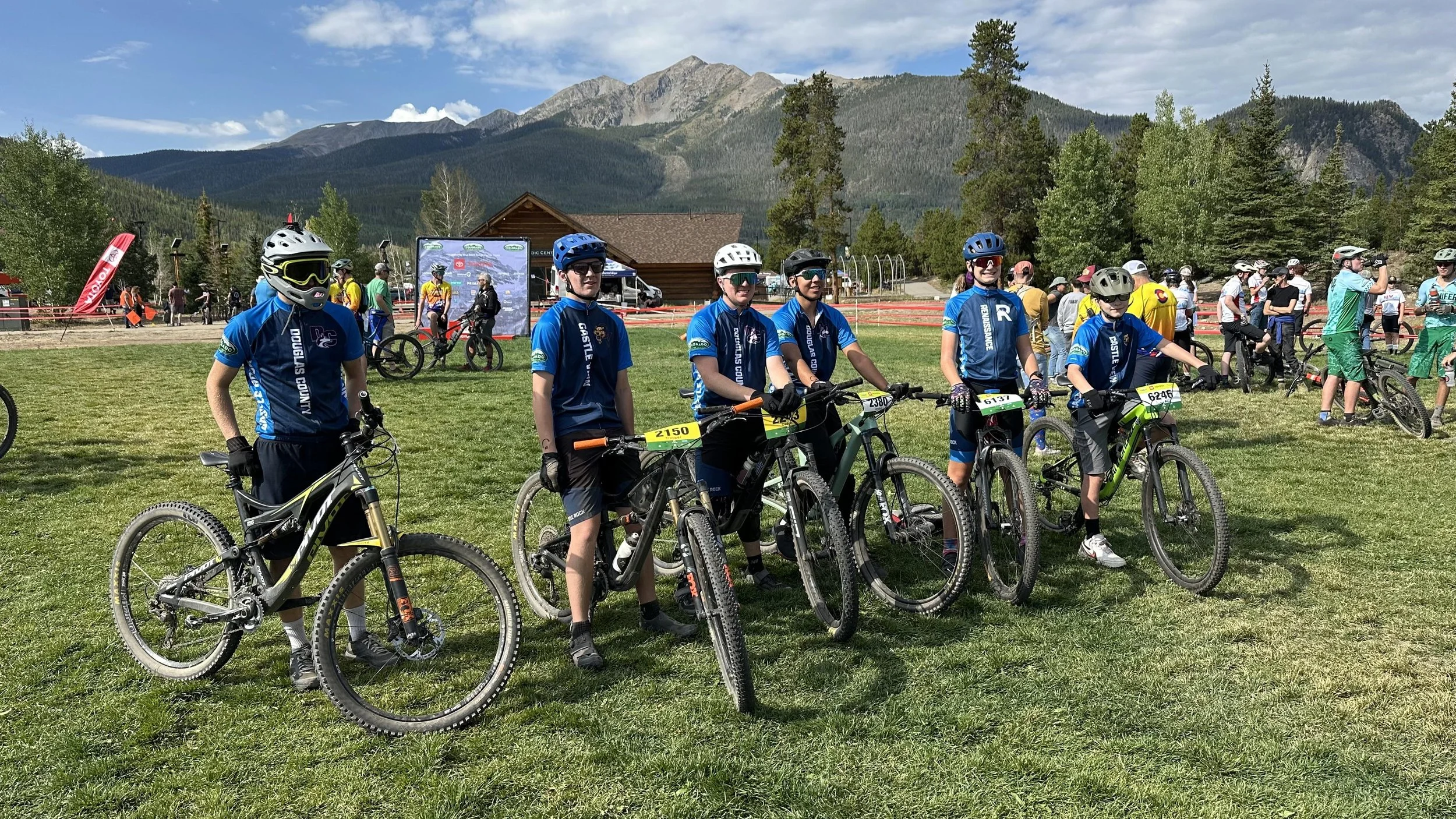 Six young cyclists in blue jerseys standing with mountain bikes on a grassy field at a mountain biking event with a mountain range and trees in the background.