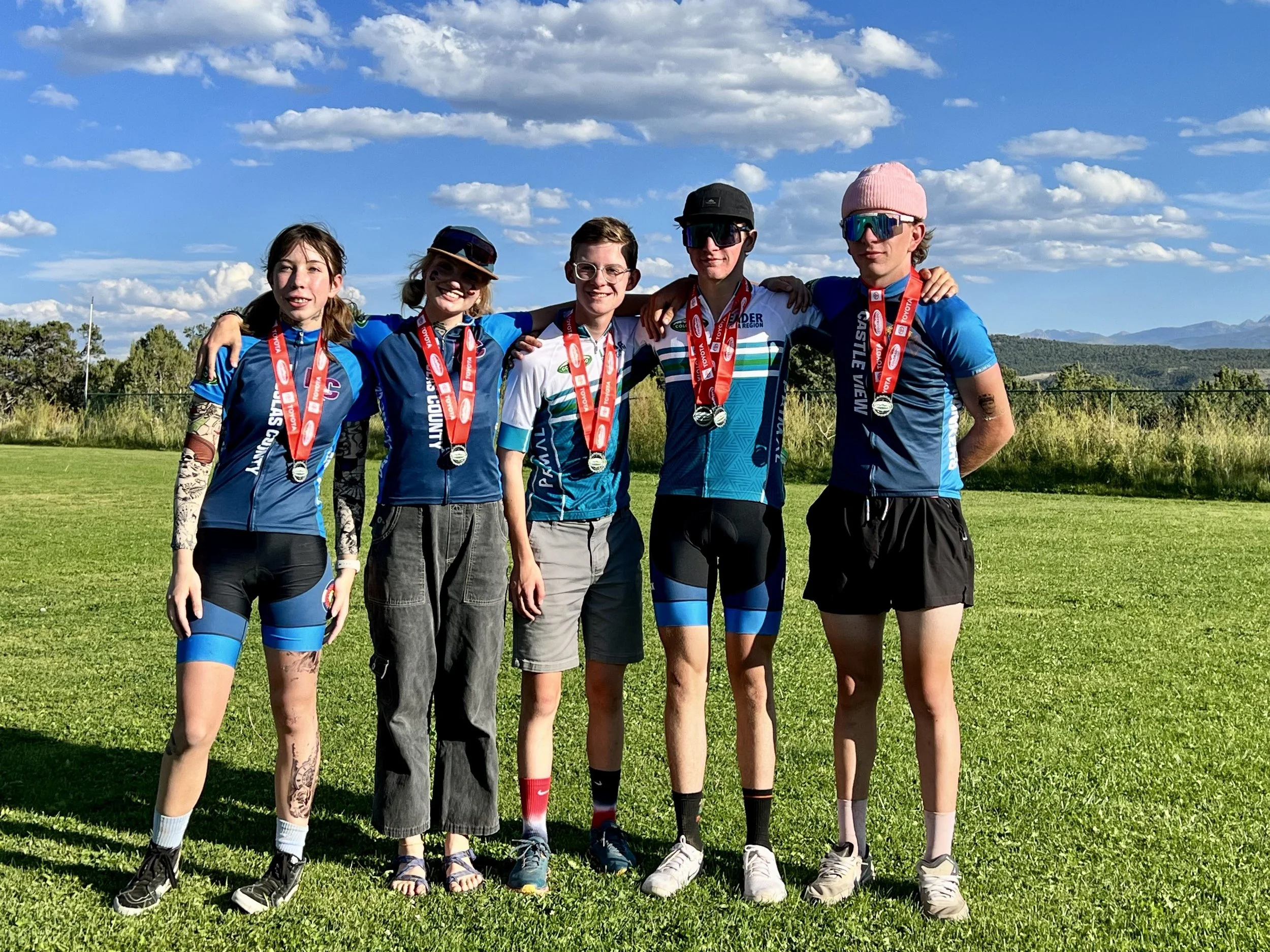 Group of five young athletes wearing medals and cycling gear standing outdoors on grass with a scenic background of blue sky, clouds, and mountains.