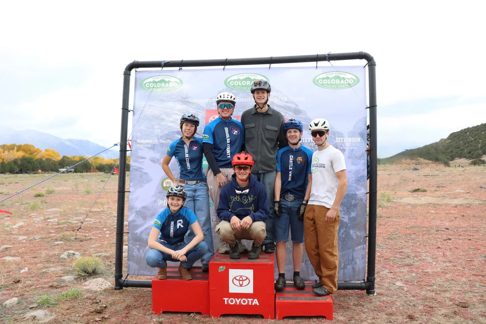 Group of six young cyclists with helmets and athletic gear, posing on a red Toyota-branded podium outdoors with a mountain landscape in the background, in front of a Colorado Cycling League banner.