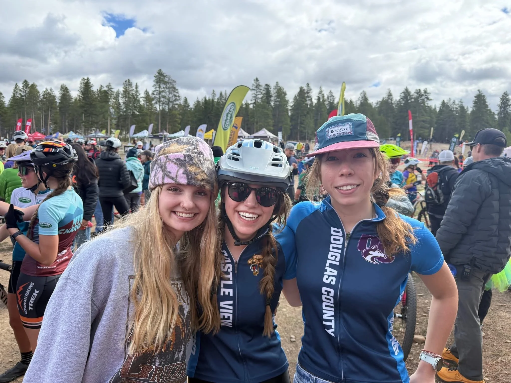 Three young women smiling outdoors at a mountain biking event, wearing cycling jerseys, helmets, and casual clothing, with many people and flags in the background.