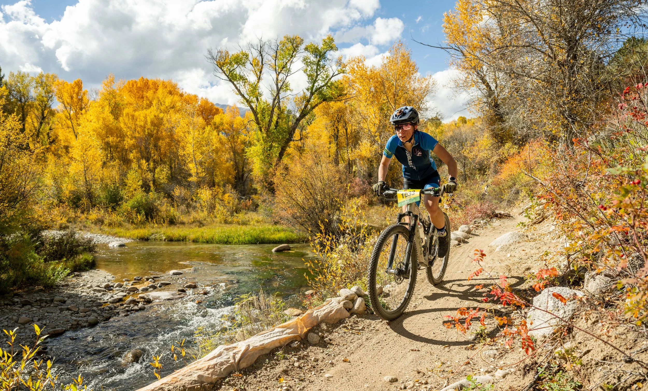 A person mountain biking on a dirt trail lined with colorful autumn foliage, next to a creek with clear water.