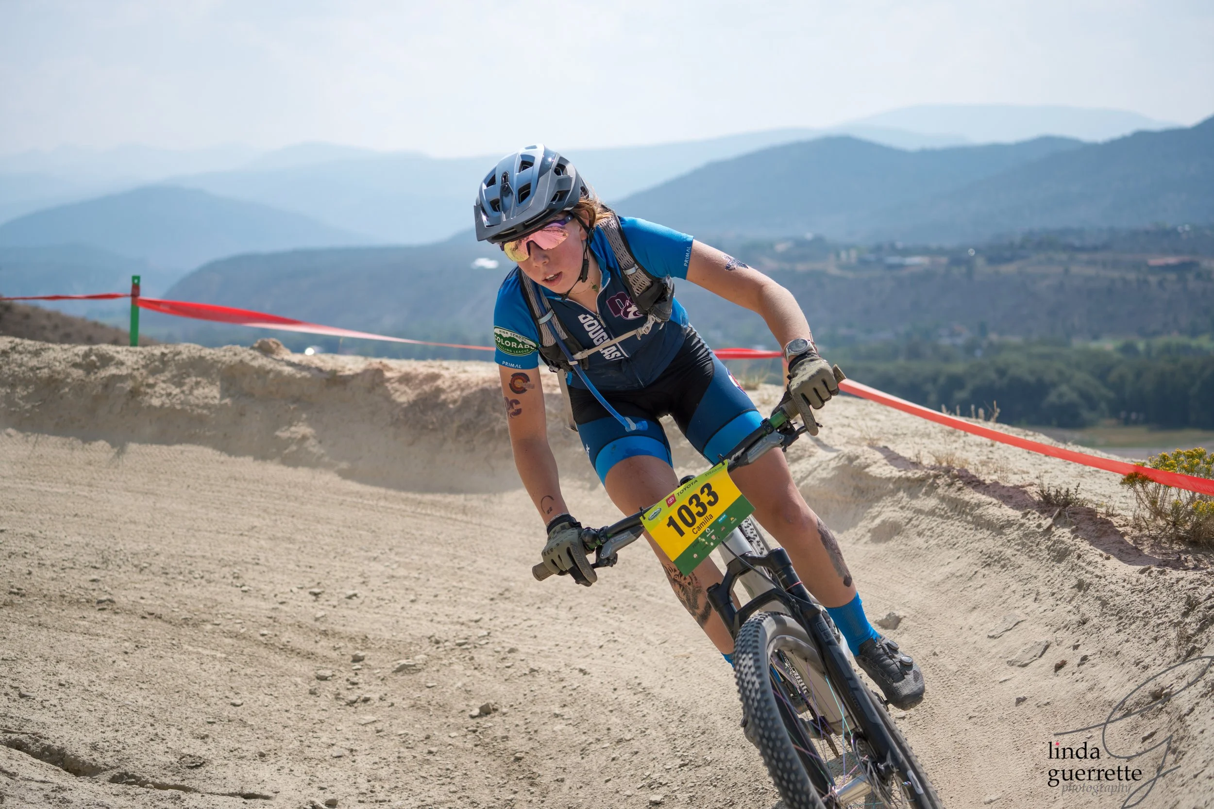 Female mountain biker wearing a blue jersey and helmet downhill biking on dusty trail with mountainous background