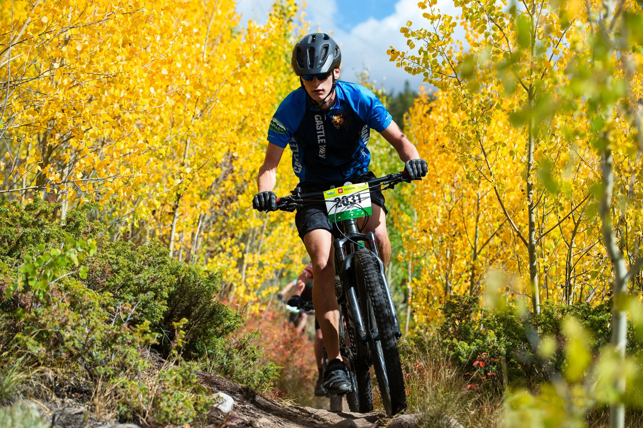 A person mountain biking on a dirt trail surrounded by yellow and green autumn trees, wearing a helmet and sunglasses.