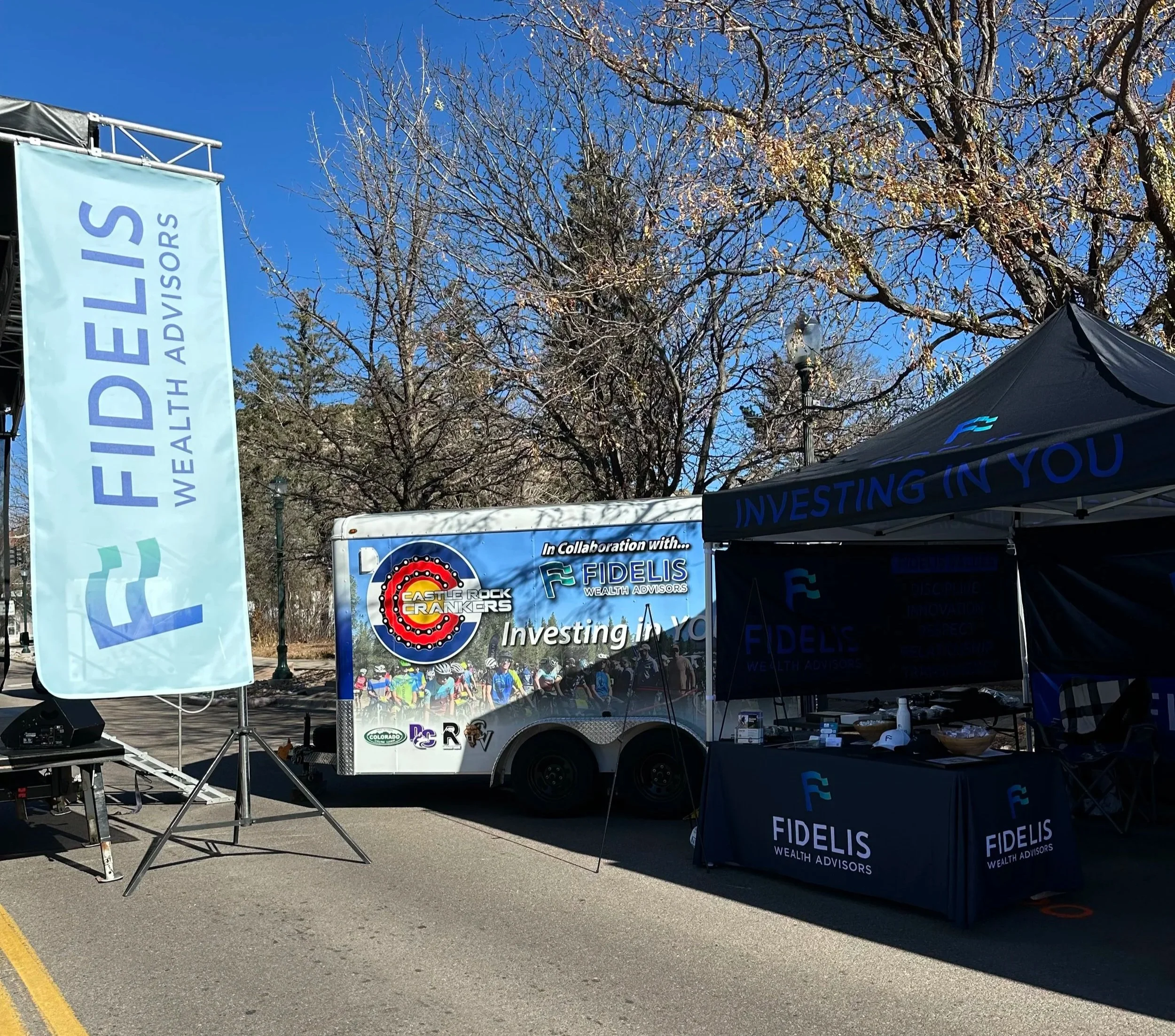 Outdoor event setup with banners and tents promoting Fidels Wealth Advisors and Castle Rock Crankers, featuring a trailer with sponsored graphics, trees with leafless branches, and a clear blue sky.