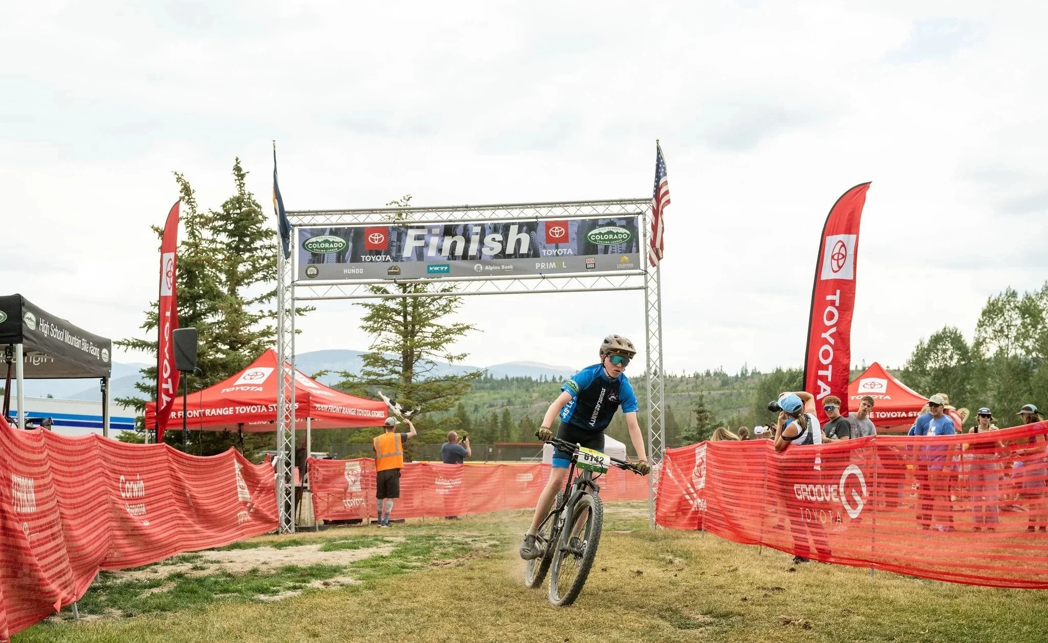 Young male cyclist in blue riding a mountain bike towards the finish line at a race event, with banners and spectators in the background.