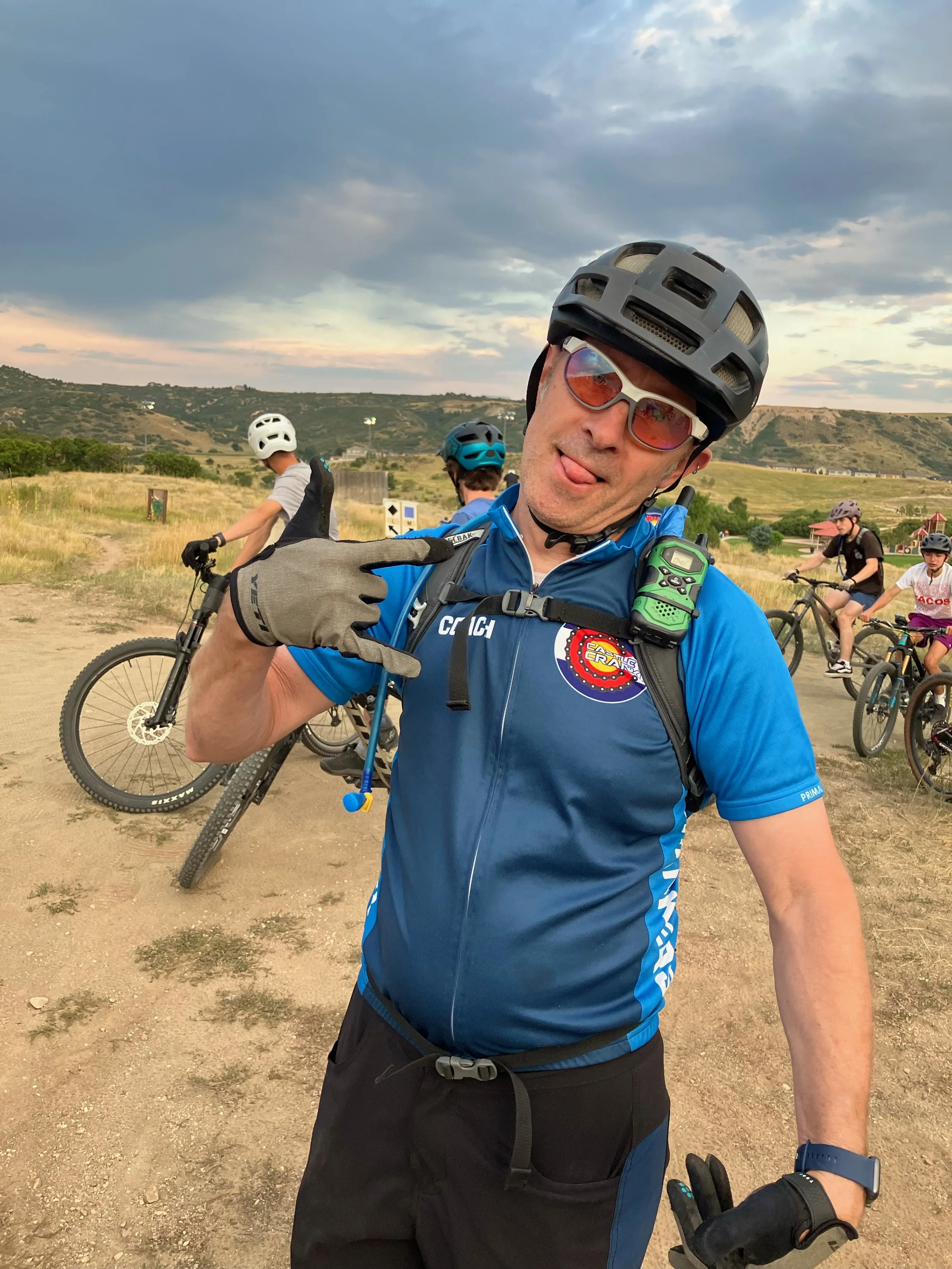 A man in cycling gear, wearing a helmet and sunglasses, standing with a group of cyclists on a dirt trail with hilly landscape in the background. The man is making a hand gesture and sticking his tongue out mischievously.
