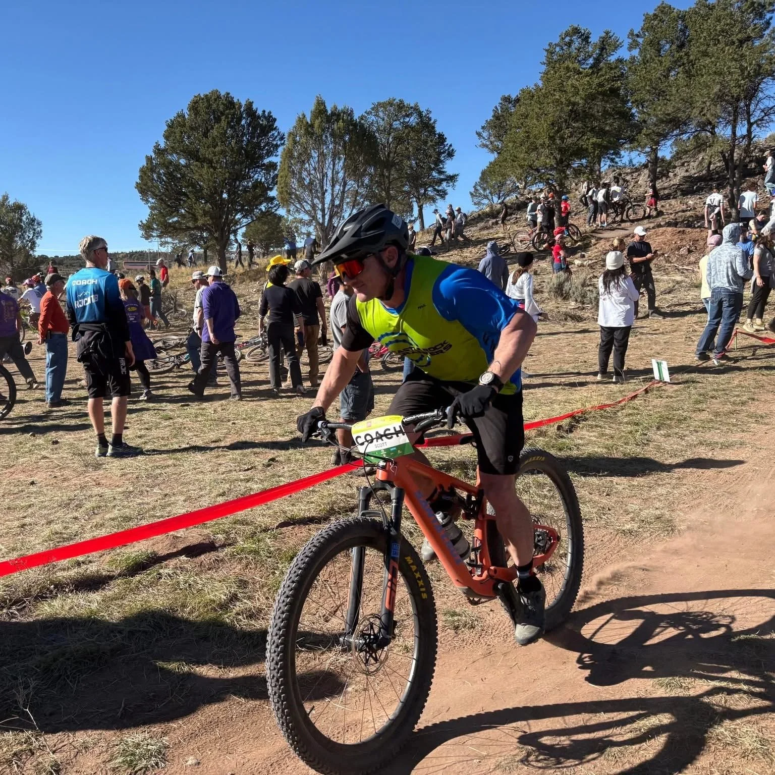 A mountain biker wearing a helmet, sunglasses, a bright yellow vest, and black gloves riding a red bike on a dirt trail during a race, with a crowd of spectators and other participants in the background on a sunny day.