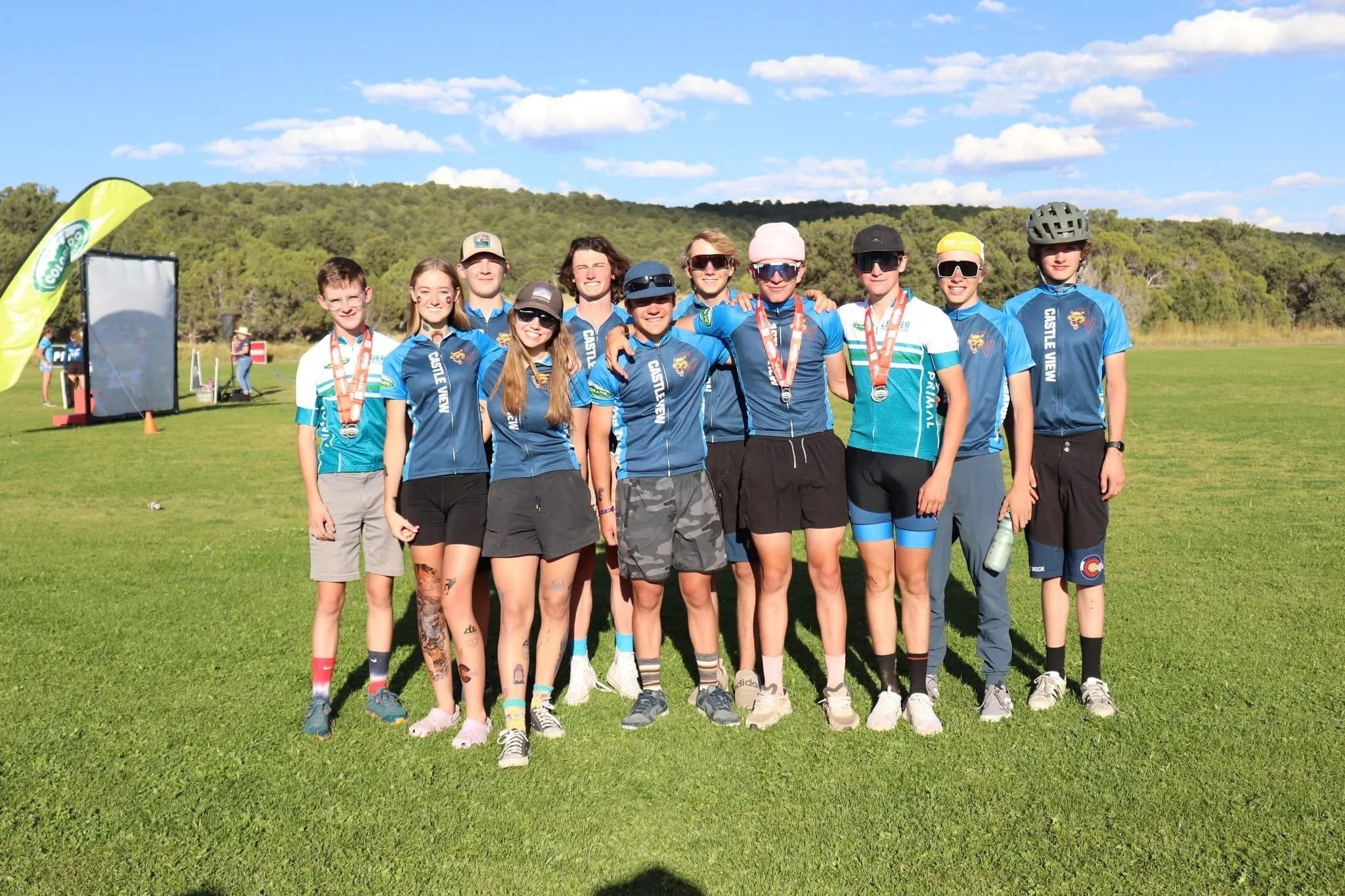 A group of ten young athletes standing on a grassy field, wearing sports uniforms and medals, smiling at the camera with arms around each other.