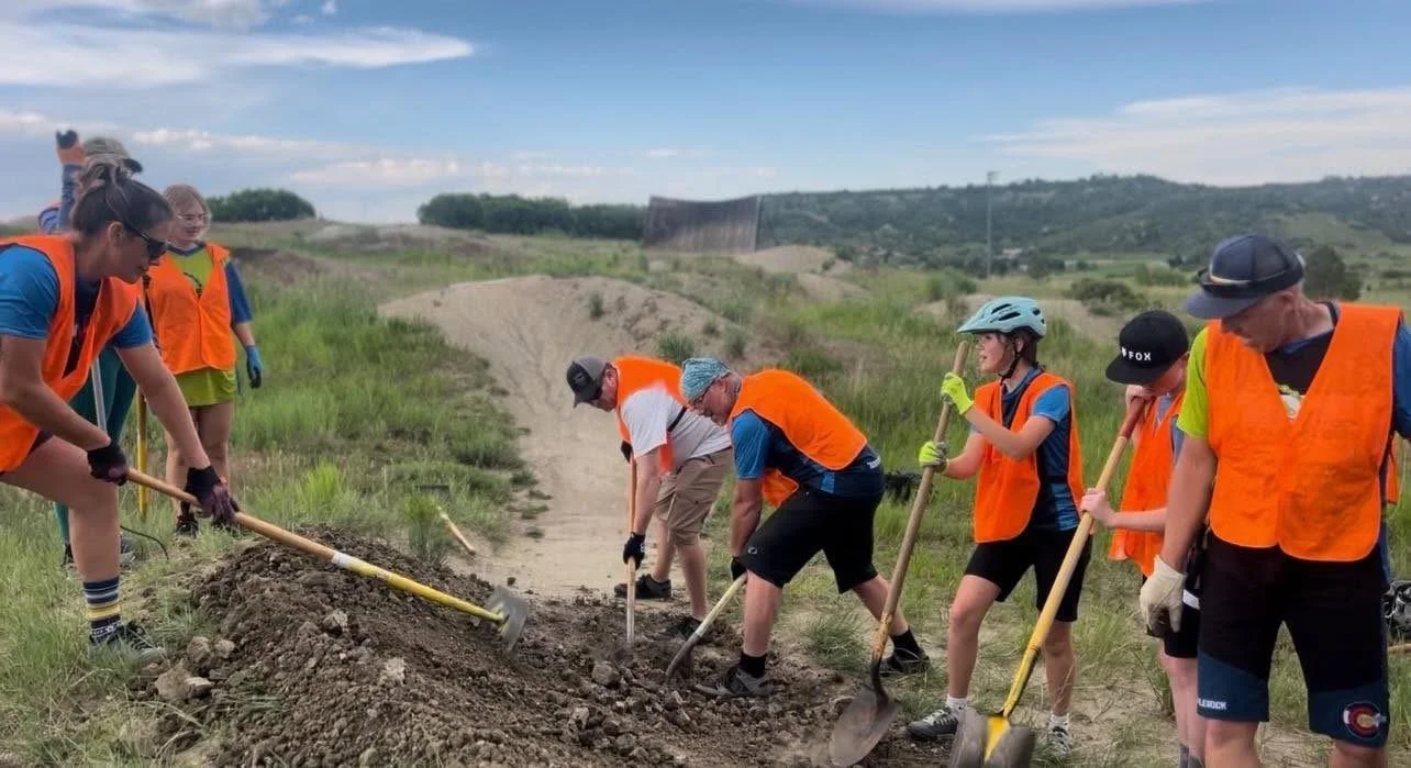 Group of people in orange safety vests and hats planting and working on a dirt trail in a rural area with hills and blue sky
