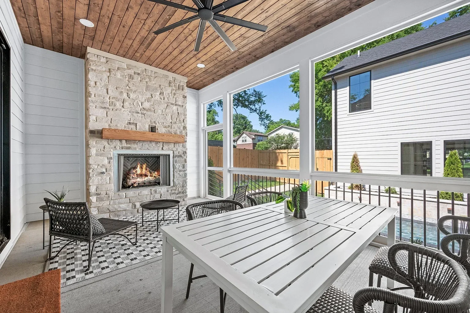 Covered outdoor patio with a white wooden dining table, woven chairs, a black and white patterned rug, a brick fireplace, large screens, and a ceiling fan, overlooking neighboring houses and trees.