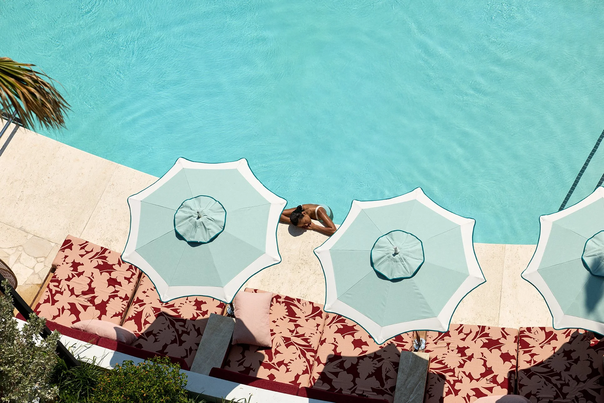 A woman relaxing by a swimming pool, sunbathing underneath beach umbrellas on a lounge area with red floral cushions.