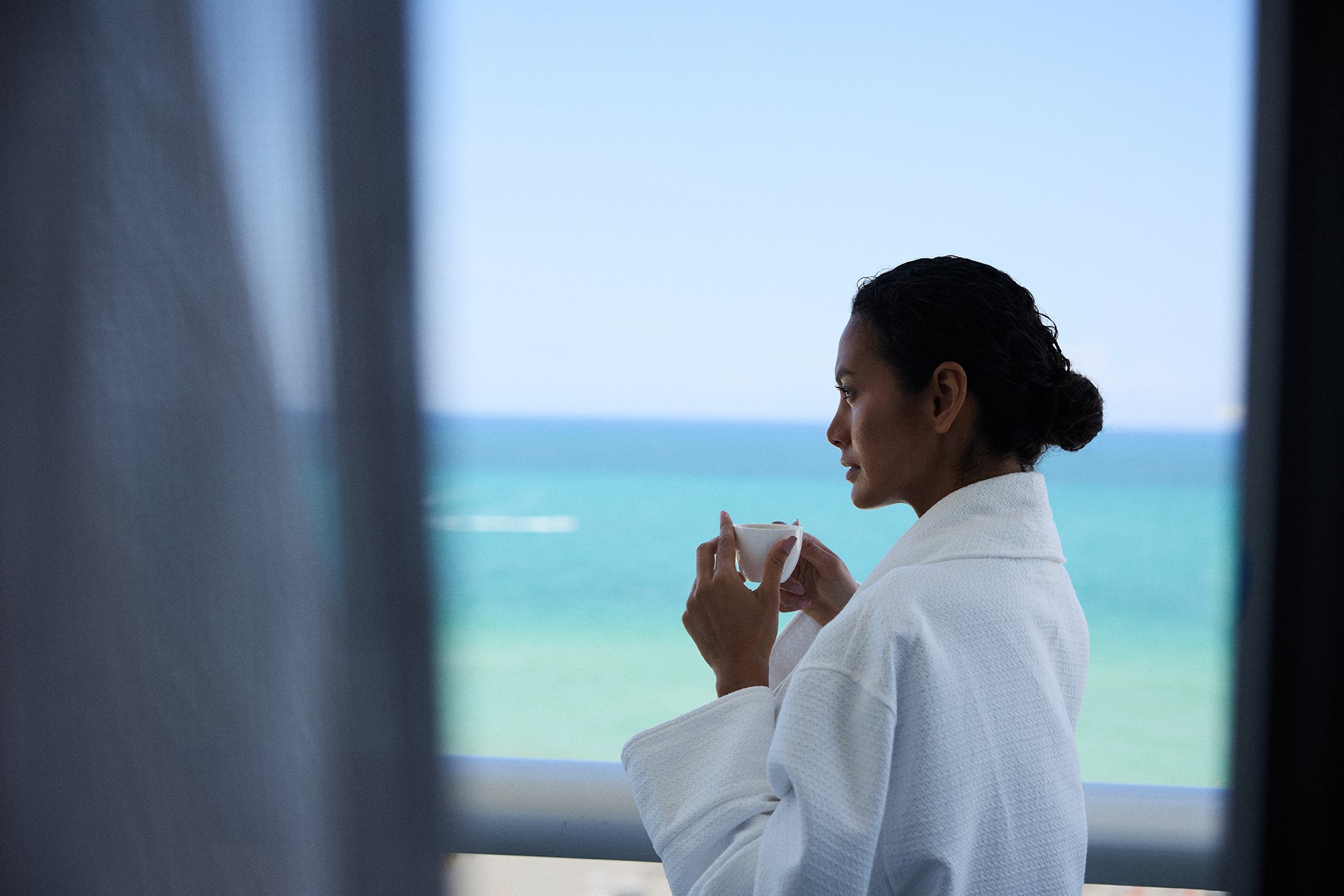 A woman in a white bathrobe holding a cup and looking out at the ocean, viewed through what appears to be a window or curtain.