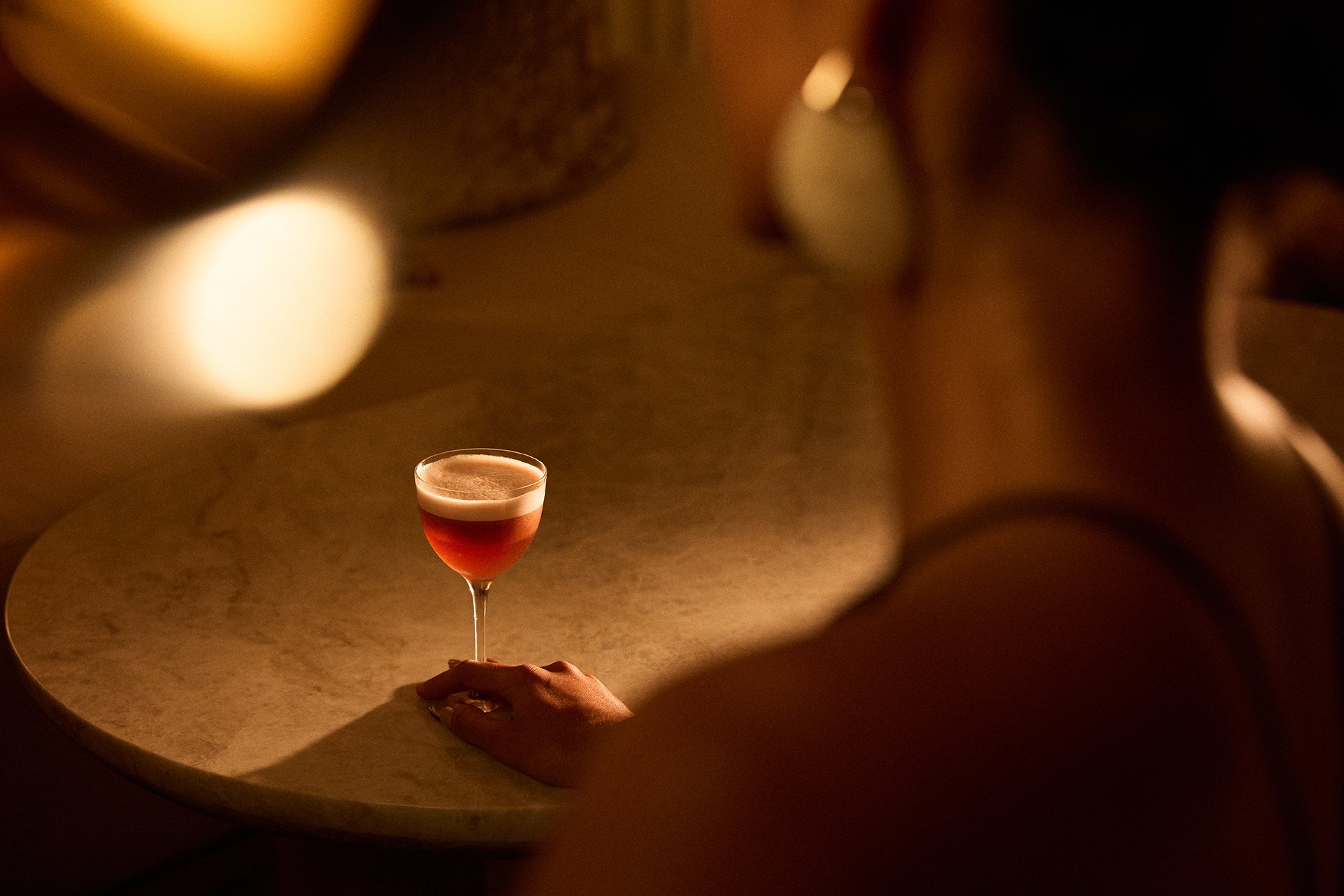 A person, partially visible, with a hand resting on a table, holding a cocktail glass filled with a red drink, seen from behind in a dimly lit setting.