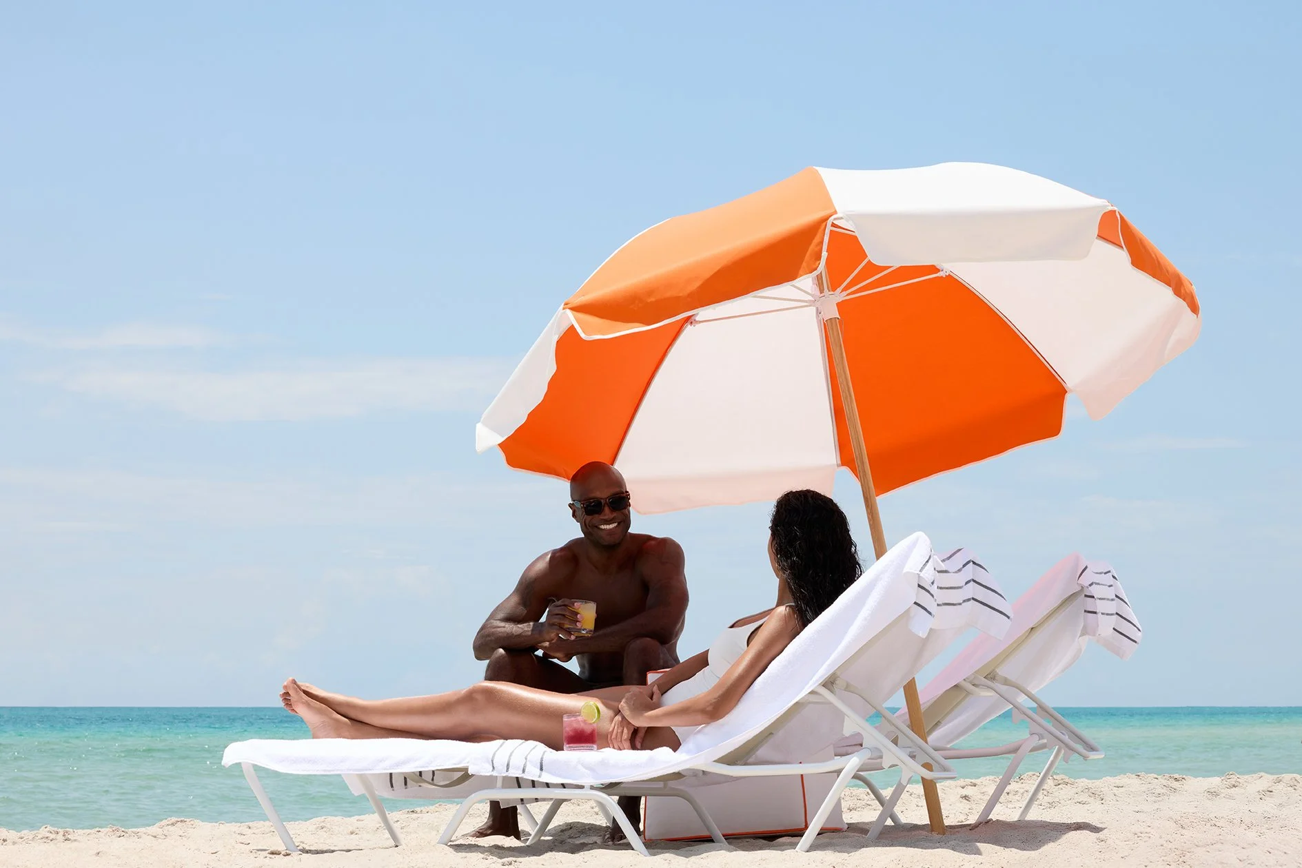 A man and a woman relaxing on beach chairs under an orange and white umbrella on a sandy beach near the ocean, with drinks on the chairs.