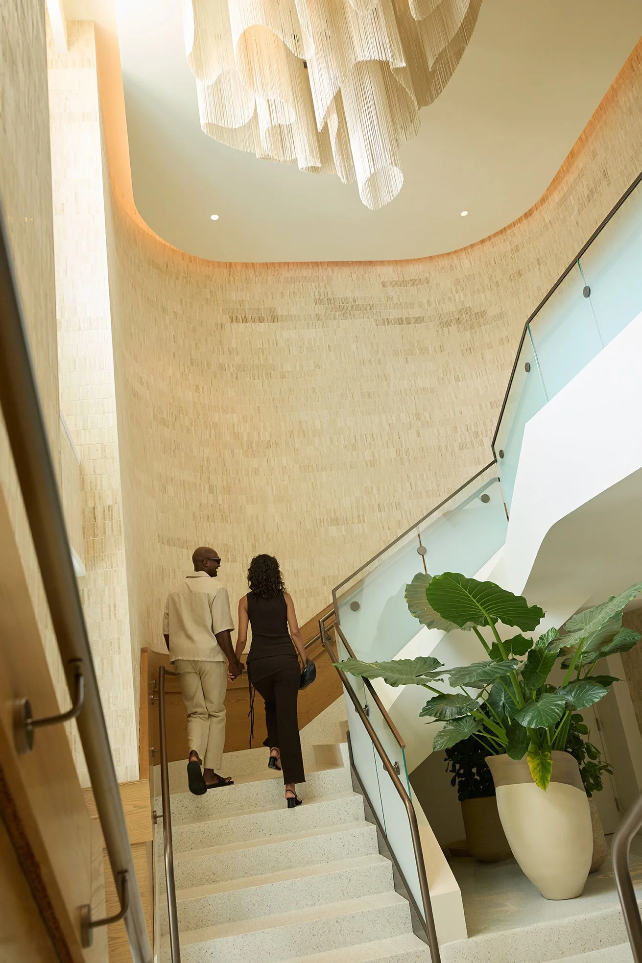 A couple walking up a staircase in a modern building, with a large green potted plant on the right and a decorative chandelier hanging from the ceiling.