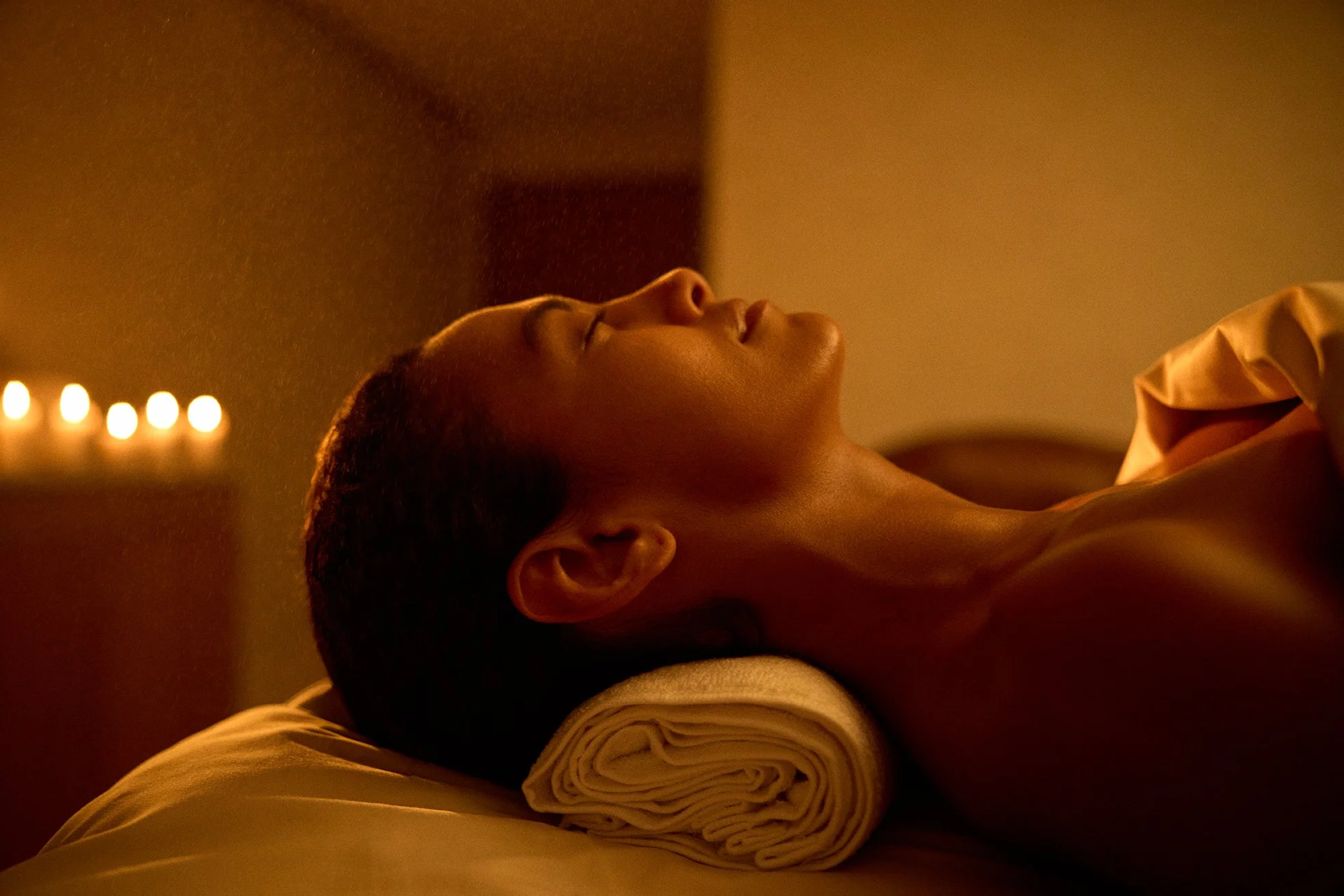A woman lying on her back with eyes closed, resting her head on a rolled towel, in a dimly lit room with candles in the background.