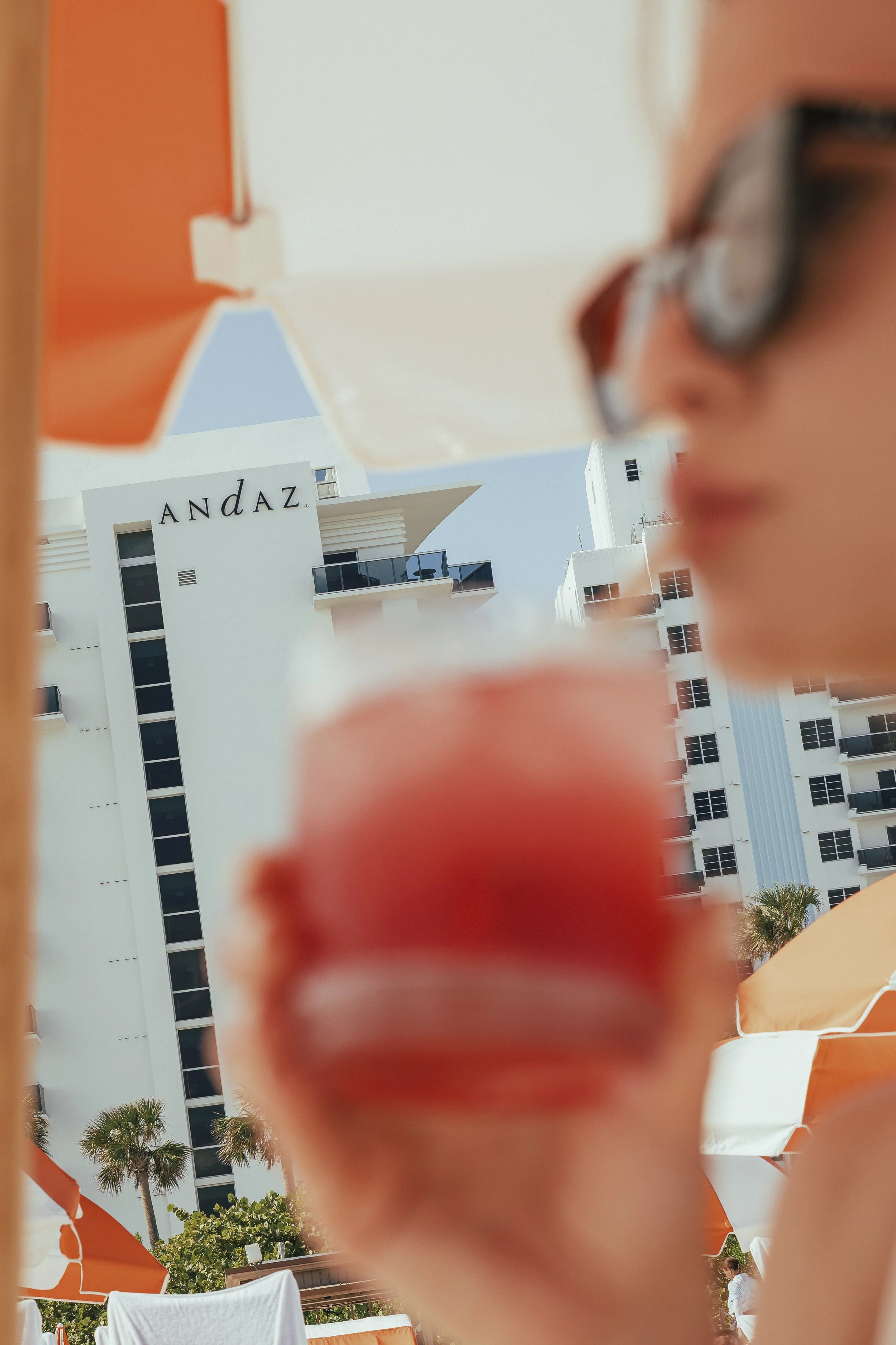 A woman with glasses and makeup in the foreground, blurred, holding a red popsicle with a hotel building labeled 'ANDAZ' and beach umbrellas in the background.