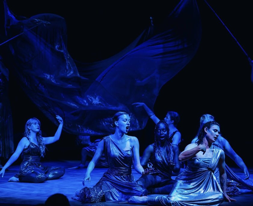 Group of women in shiny blue costumes performing a dance on a dark stage with flowing fabric.