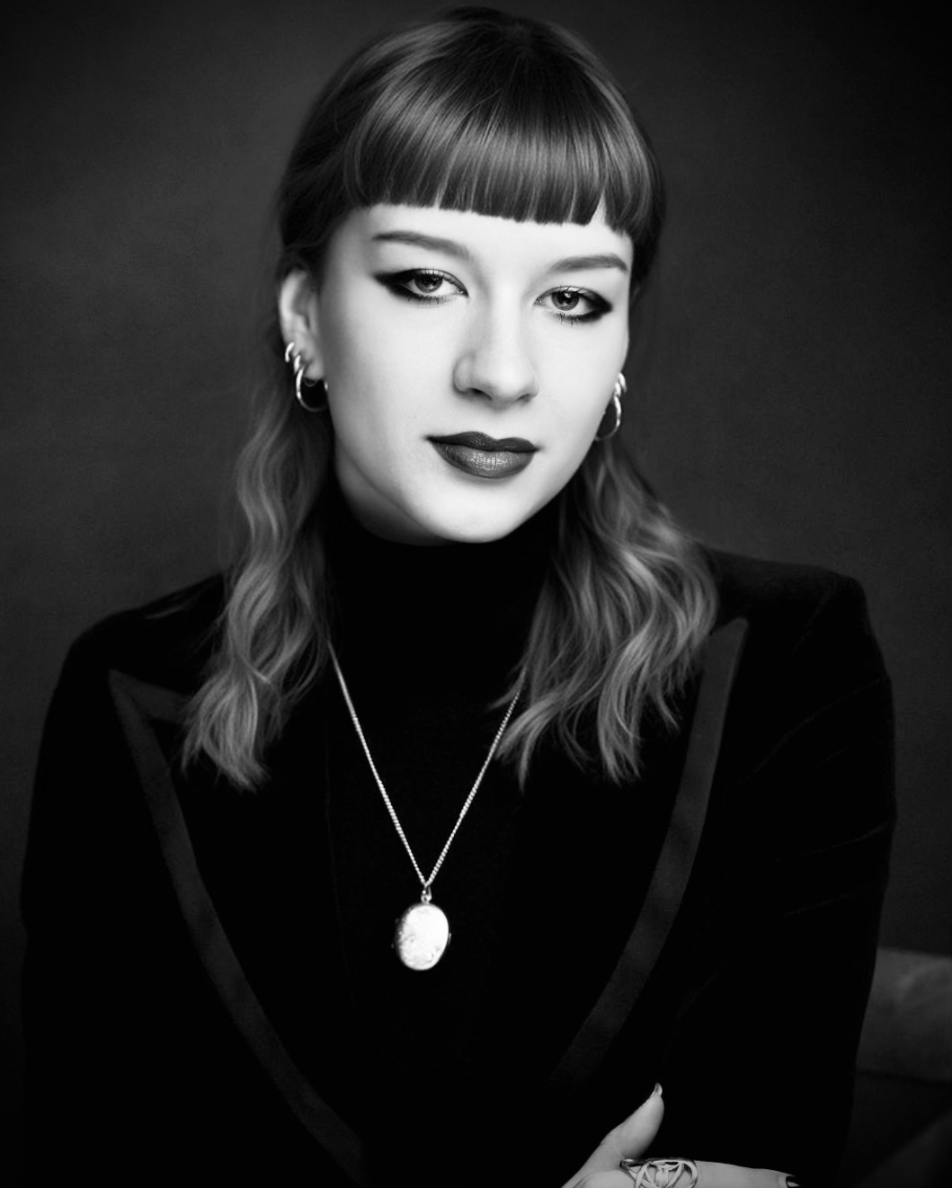 Black and white portrait of a woman with shoulder-length wavy hair, wearing earrings, a necklace, and a dark top, looking directly at the camera.